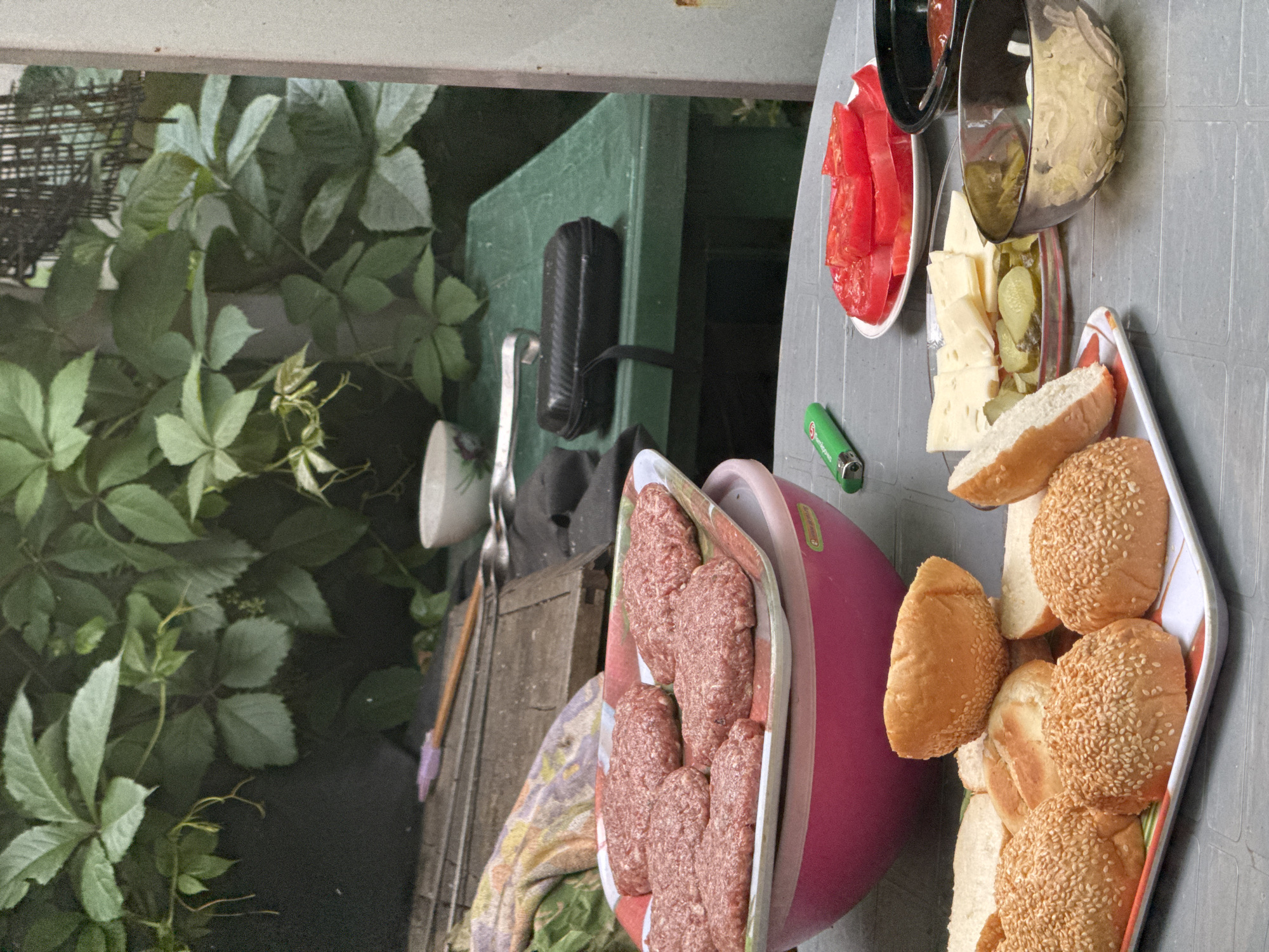 The image depicts an outdoor scene, possibly a backyard barbecue, with various food items arranged on a grey, textured table. In the foreground, a tray holds several hamburger buns, some sprinkled with sesame seeds. To the right of the buns, a plate contains sliced tomatoes, and next to it, a small bowl holds sliced pickles and cheese. Further to the right, a black bowl contains a red condiment, likely ketchup, and another bowl contains what appears to be shredded cabbage or coleslaw. A bright green lighter rests on the table near the plate of tomatoes. In the background, a pink bowl is visible, holding numerous uncooked hamburger patties. Behind the food, a wooden plank lies on a green surface, with some gardening tools and a patterned cloth. Lush green foliage hangs in the upper portion of the image. The overall setting suggests a casual meal preparation. The location is identified as Pribrezhnyi, Russia.