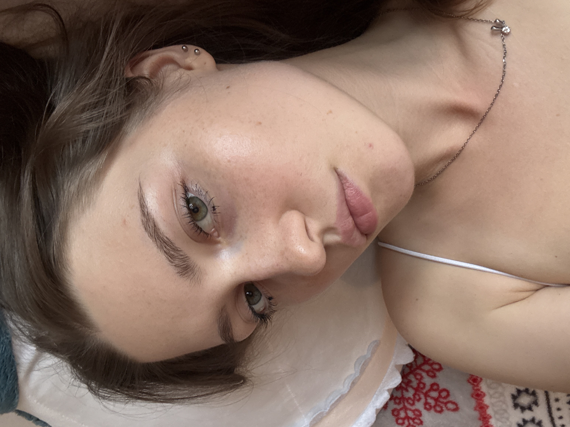 A close-up shot shows a young woman with light skin and brown hair lying down, looking upwards and to the right. She has green eyes, light freckles across her nose and cheeks, and subtle makeup. Her hair is slightly messy and falls around her face. She is wearing a white tank top and a silver necklace with a small charm. The background is a soft, textured surface, possibly a pillow or bedding, with a decorative embroidered pattern in red and white visible on the left side. The overall lighting is soft and natural. The image is a selfie, taken from a high angle. There is no information about the location in Saint Petersburg, Russia.