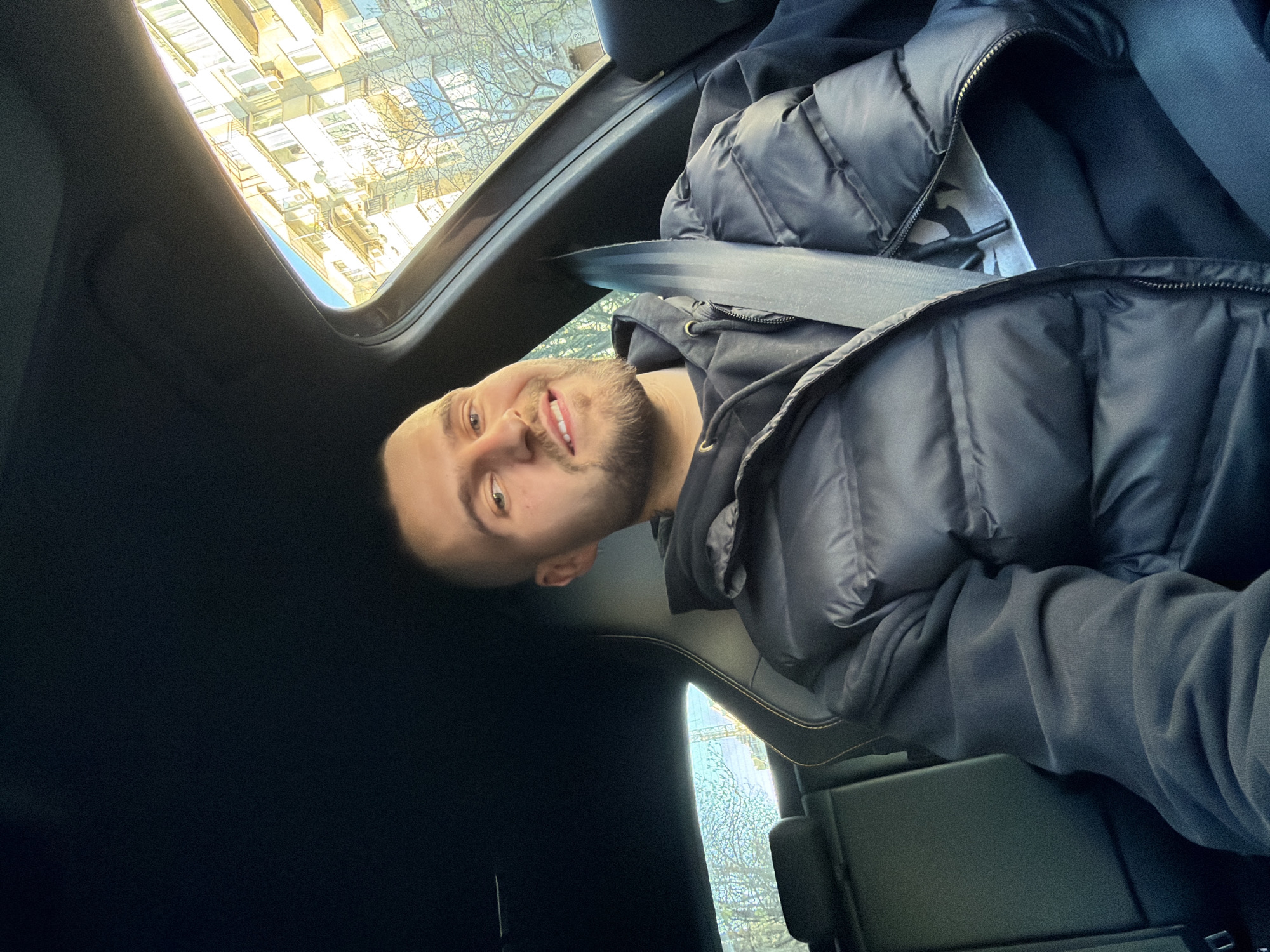 A man with a beard, wearing a black puffy jacket and a seatbelt, is sitting in a car. He is smiling at the camera. The car has a sunroof that is partially open, revealing a view of buildings and trees outside. The location appears to be in Sofia, Bulgaria, based on the architectural style of the buildings visible through the sunroof. The interior of the car is black leather.