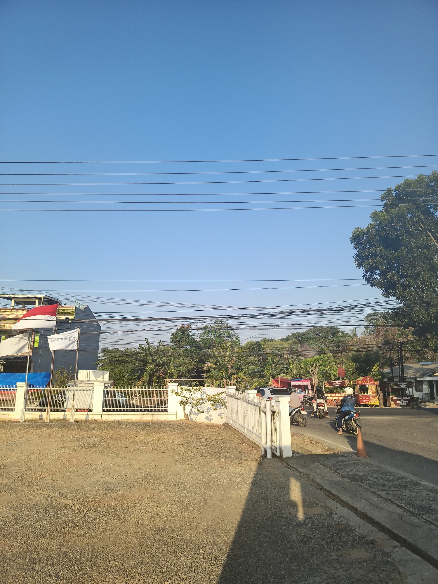 The image depicts a street scene in Pangkalpinang, Indonesia, on a clear, sunny day. In the foreground, there is a gravel lot with a shadow cast by a fence and a white gate. Beyond the fence, a paved road runs horizontally across the frame, with several motorbikes visible. One motorbike, ridden by a person wearing a blue jacket and white helmet, is stopped near an orange traffic cone. To the left of the road, there is a building with flags, including what appears to be the Indonesian flag. Further down the road, various food stalls or small businesses are set up. The background is filled with lush green trees and utility wires crisscross the clear blue sky. The overall impression is one of a typical urban streetscape in a tropical climate.