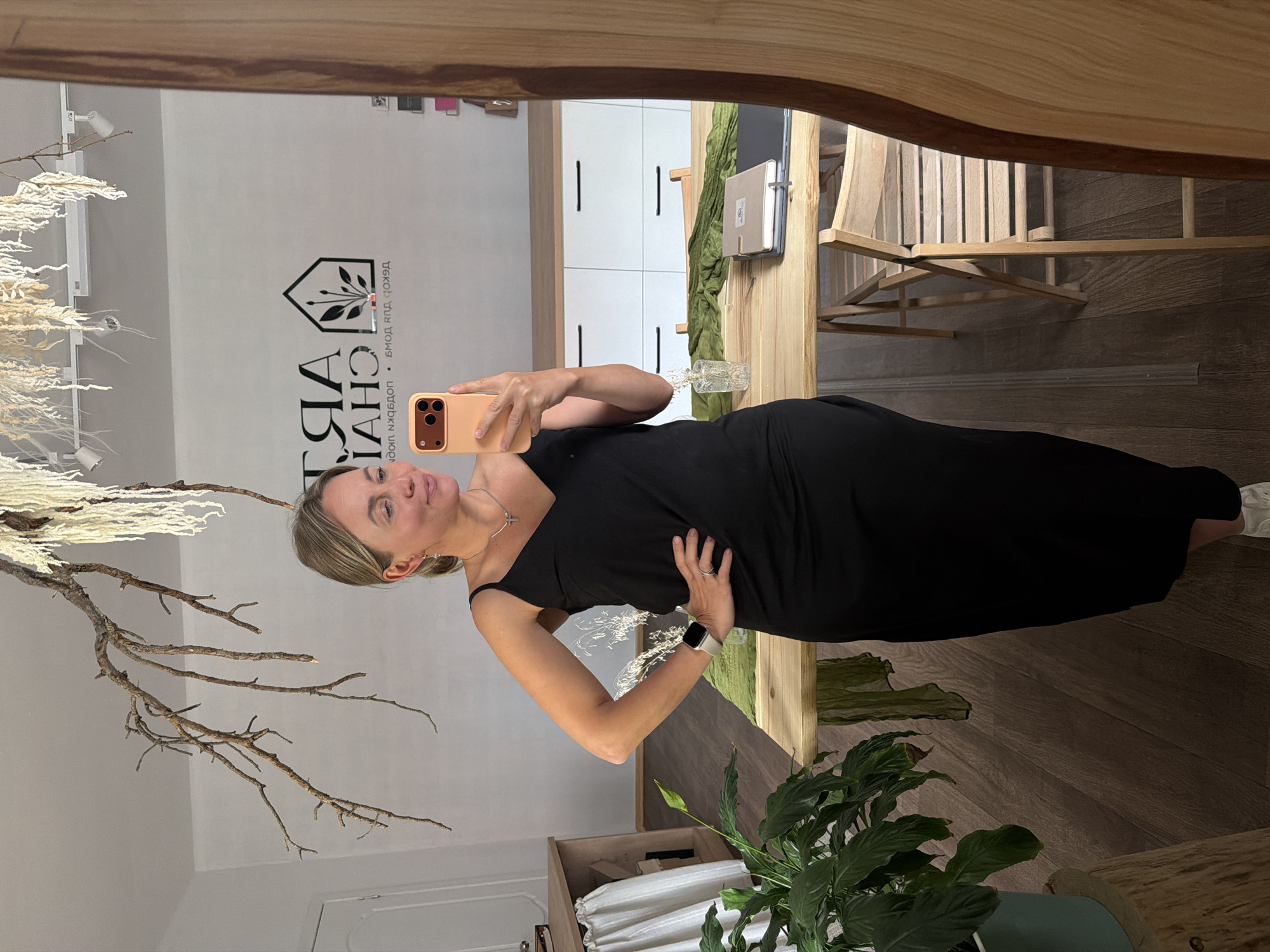 A woman in a black dress stands in a room and takes a mirror selfie. She has blonde hair pulled back into a ponytail and is wearing a silver necklace with a cross pendant and a silver watch on her left wrist. She is holding an orange smartphone in her right hand. The room features wooden floors, a wooden table with chairs, and white cabinetry. A large plant sits in a pot on a wooden stump in the foreground. A branch with dried flowers hangs from the ceiling. In the background, a logo is visible on the wall, featuring stylized text and a house with a plant inside. The location is identified as Chaikovsky, Russia.