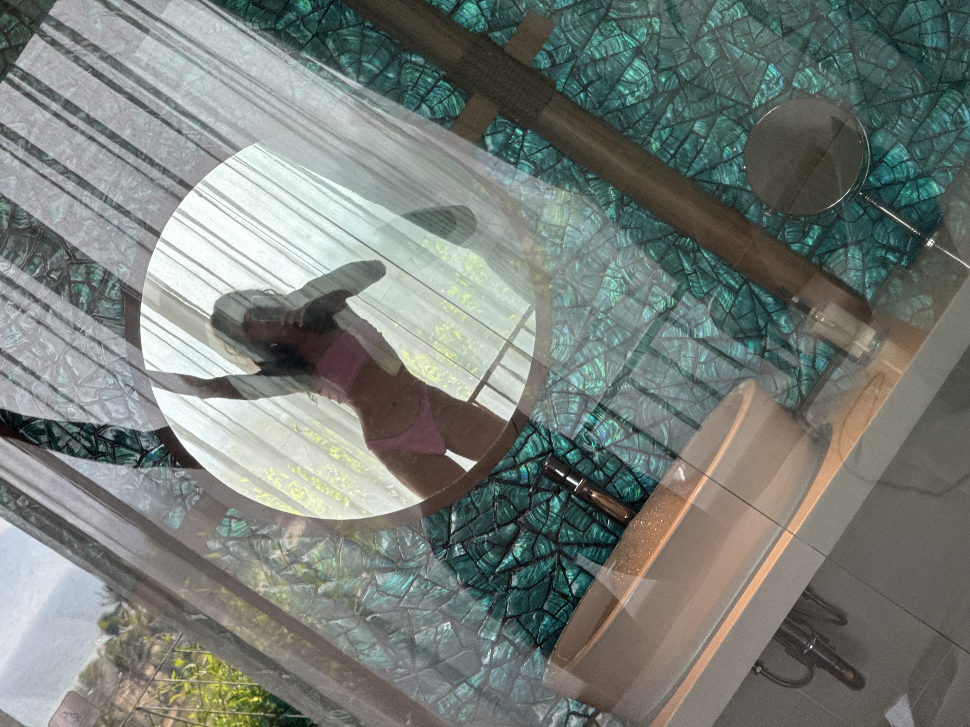 The image shows a woman in a pink bikini, viewed through what appears to be a circular window. Outside the window is a landscape with greenery. The location appears to be a bathroom in Đà Nẵng, Vietnam, as indicated by the distinctive patterned tile work on the bathroom surfaces, including around the tub and possibly the walls or floor. A vanity mirror and other bathroom fixtures are visible. The image is likely taken through a glass panel or a reflection.