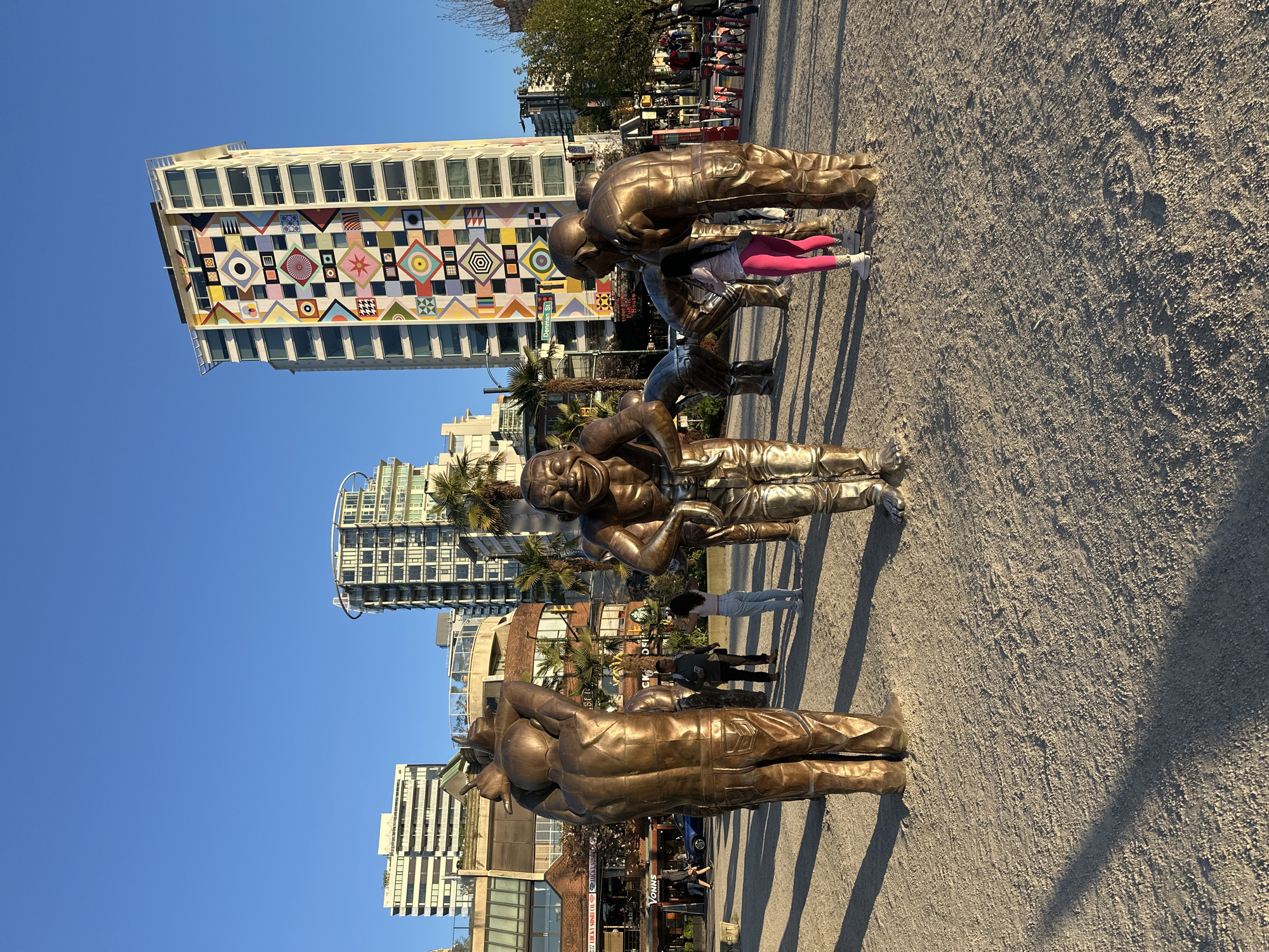 This outdoor scene captures a vibrant public space in Vancouver, Canada, featuring a prominent bronze sculpture installation against an urban backdrop under a clear blue sky. Five large, stylized bronze statues of human figures are arranged linearly on a light-colored, textured ground. The statues depict various poses; in the foreground, one figure points, followed by another with a laughing expression, and two figures bent forward, with a final figure standing behind them. Long shadows cast by the statues suggest either early morning or late afternoon sun.
Several real people are present in the scene. One person wearing bright pink pants is crouched near one of the bronze statues, while others stand further back, observing or walking.
The background is dominated by modern city architecture. A striking multi-story building features a large, colorful facade composed of geometric mosaic patterns in various shapes and hues, including squares, circles, and triangles. Other high-rise buildings with glass and concrete exteriors are also visible, some with lush palm trees interspersed.
Visible text includes a street sign reading "Denman St" and a building sign with "VONNS". The overall atmosphere is bright and inviting, showcasing public art integrated into an urban environment.