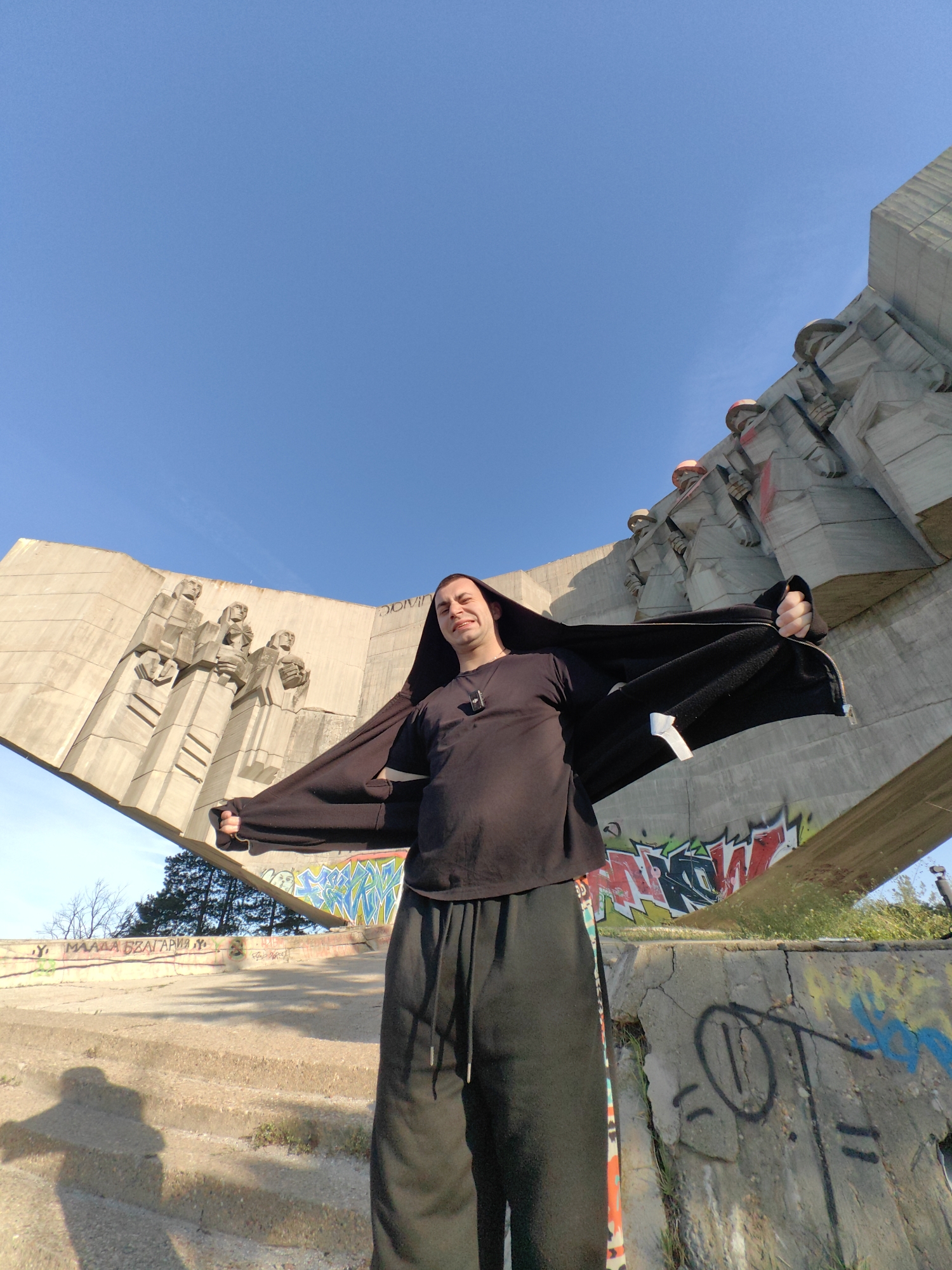 The image shows a man standing in front of the Monument of the Bulgarian-Soviet Friendship in Varna, Bulgaria. He is wearing a black t-shirt and black pants, and holding a black jacket open wide, with the hood over his head. The monument is a large concrete structure with bas-relief sculptures on its surfaces, showing depictions of people. There is graffiti on the lower parts of the monument and on the surrounding concrete structures. The sky is clear and blue. The man is looking at the camera and smiling.