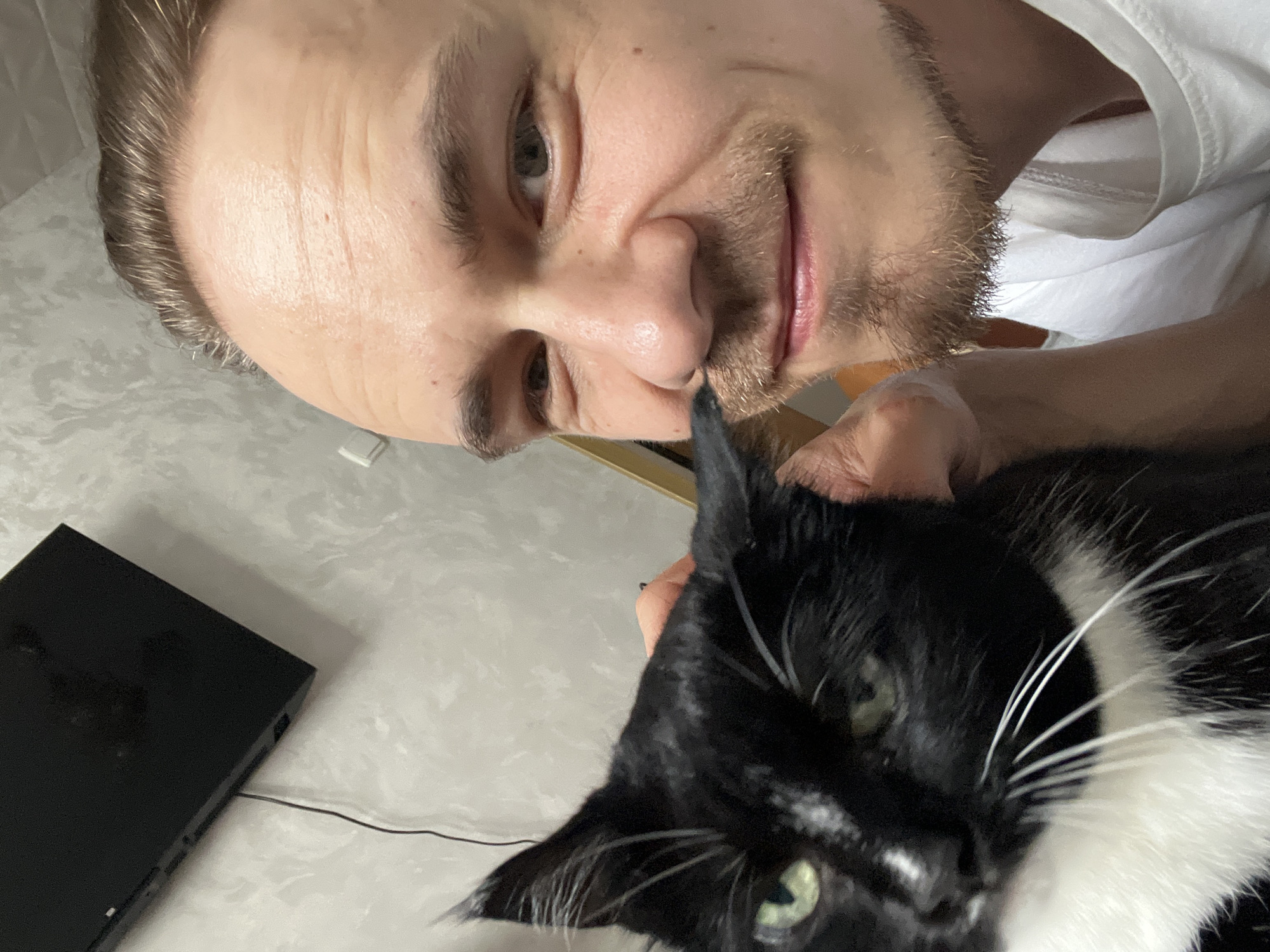 A close-up selfie taken indoors shows a person with light skin, a receding hairline, and light brown hair. They have a light mustache and stubble, and are smiling with their mouth slightly open. Their left arm is visible, holding a black and white cat that is looking towards the camera with green eyes. The background is a lightly textured wall and a black object, possibly a television, mounted on it. A wire is also visible. The image appears to be a casual, personal photograph, not indicative of any specific public location within Moscow, Russia.