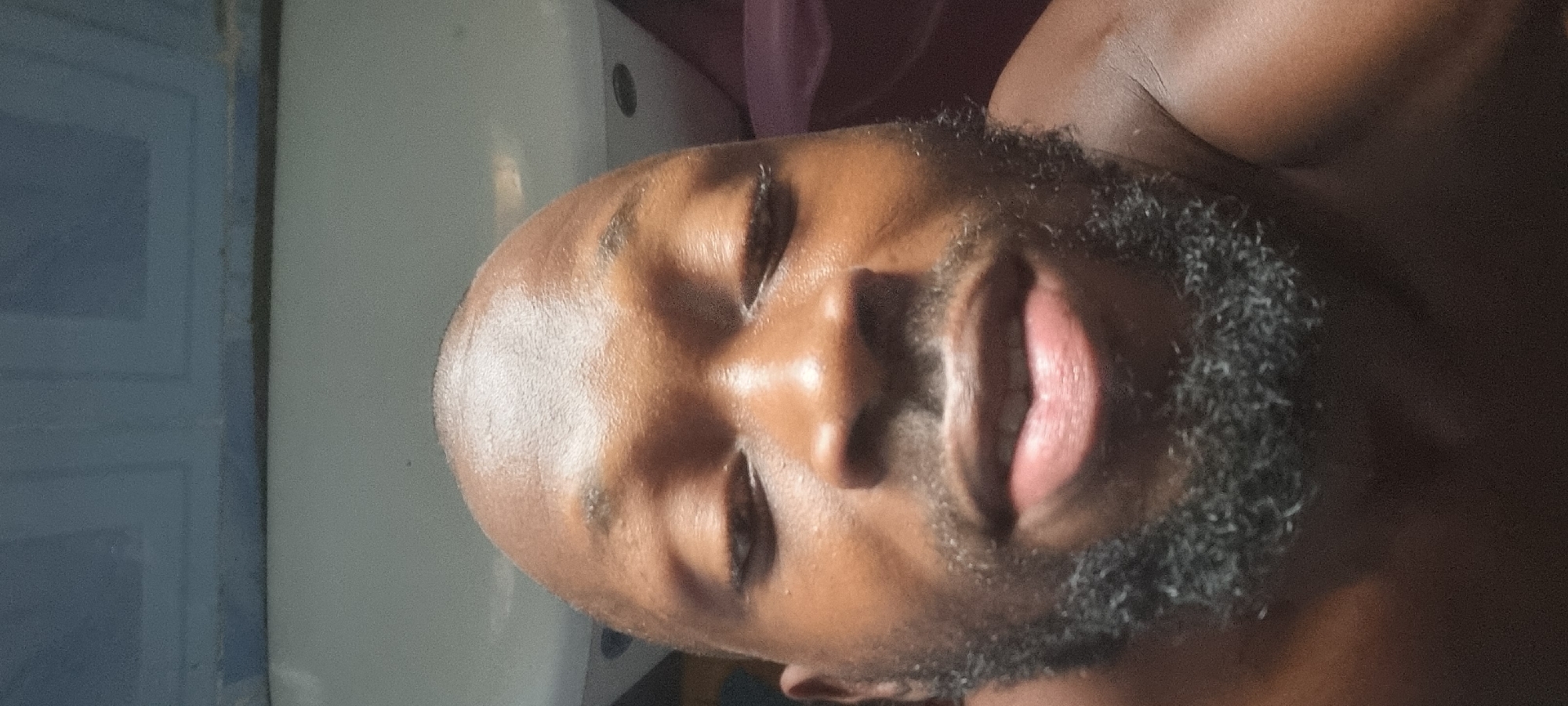 A close-up image shows a dark-skinned person with a bald head and a full, dark beard, lying down with their eyes closed. The skin appears shiny, possibly due to sweat or oil. The background is blurry, but includes a white or light-colored surface and a hint of what might be purple fabric. Based on the prompt's request for a location, the individual is presumably located somewhere in Lagos, Nigeria.