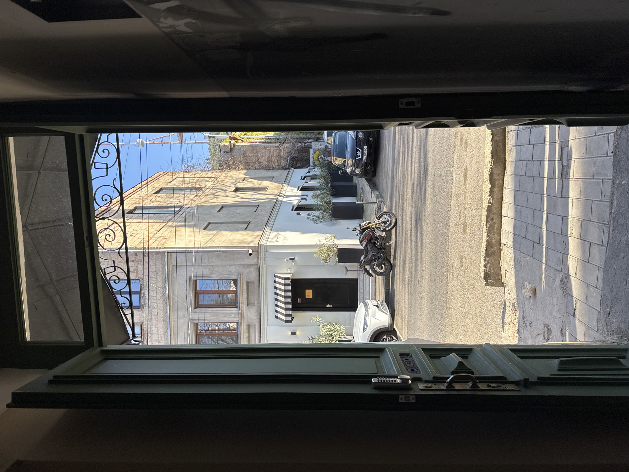 View from inside a building out onto a street in Tbilisi, Georgia. An open green door with ornate ironwork partially frames the scene. The street features parked cars, a motorcycle, and a building with a black and white striped awning. The building is adjacent to a brick building. The street is paved, and a sidewalk is visible on the right side. The sky is blue and mostly clear.