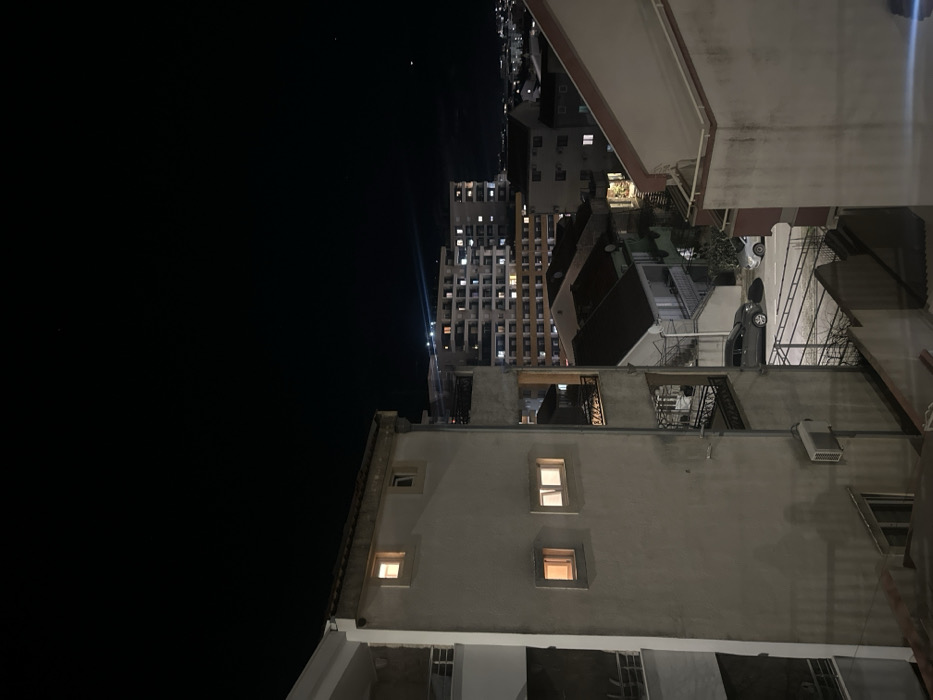 A night scene features buildings in an urban setting. Some buildings have illuminated windows, contrasting with the dark sky. The composition is taken at an angle, showcasing rooftops, balconies, and streets. A tall building with numerous lit windows is visible in the distance. A vehicle is parked on one of the streets. The foreground includes buildings with a rough, textured exterior.