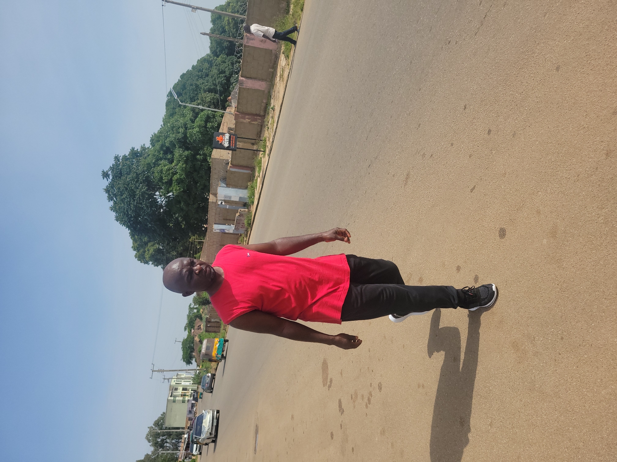 A bald Black man wearing a red sleeveless shirt and black pants walks on the right side of a paved road. He is wearing black and white athletic shoes. The man is in the foreground of the image, which is a street scene in Jos, Nigeria. In the background, there are cars, buildings, and trees. A sign for "GYM" is visible on a building to the right of the road. Another man in a white shirt and dark pants is walking away from the camera on the far right side of the road. The sky is clear and blue.