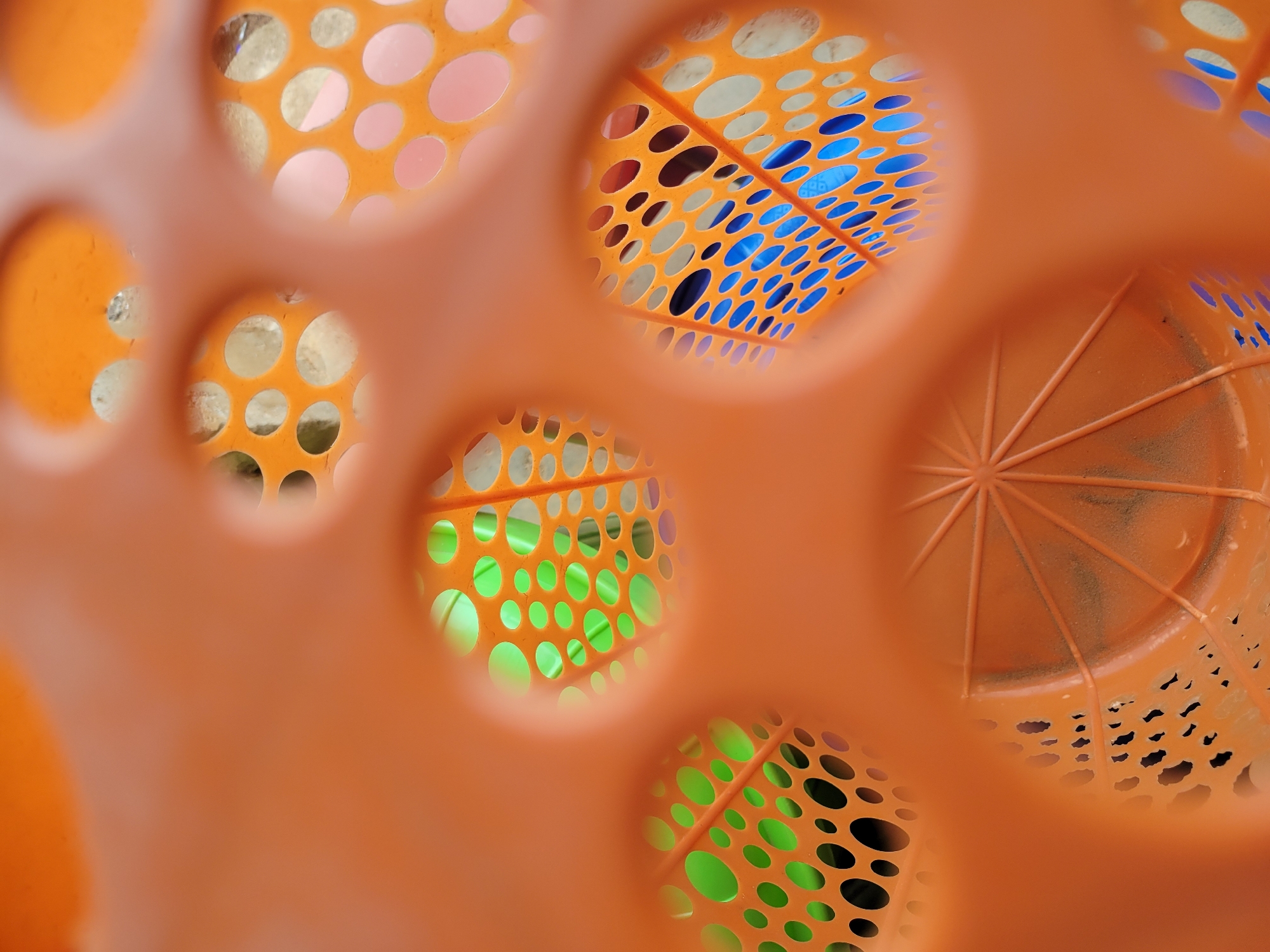 The image shows a close-up view through the round holes of an orange plastic object, likely a basket or container. The holes reveal different colored objects inside: pale yellow/beige, blue, and green. The orange plastic has a pattern of small circular perforations. In the lower right, a portion of the orange plastic's bottom is visible, showing a star-shaped pattern and additional perforations. The location and city are unknown.