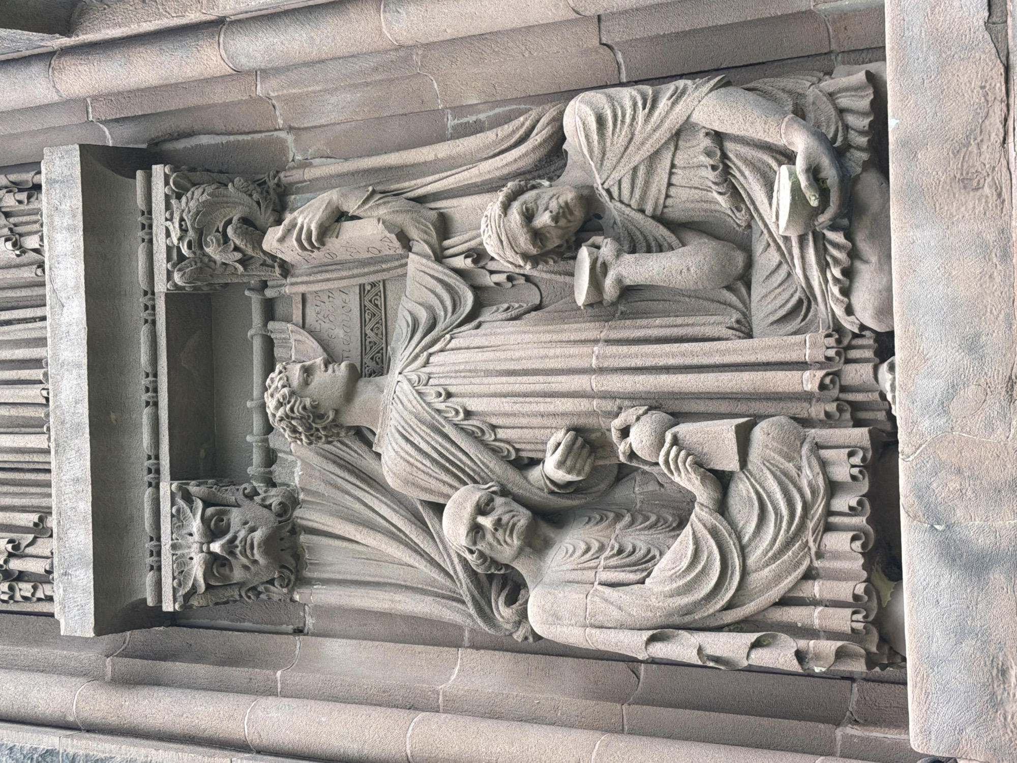 The image shows a stone sculpture on the exterior of the Princeton University Chapel, located in Princeton, New Jersey, United States of America. The sculpture depicts three male figures in classical robes. The central figure is standing and holds a book or tablet in his right hand. Flanking him are two kneeling figures. The figure on the left holds a container or vessel in his hands, and the figure on the right holds a bowl to his face. Above the central figure's head, on either side of the architectural element, are carved gargoyle-like faces. The sculpture is made of weathered stone and is integrated into the gothic architecture of the chapel.