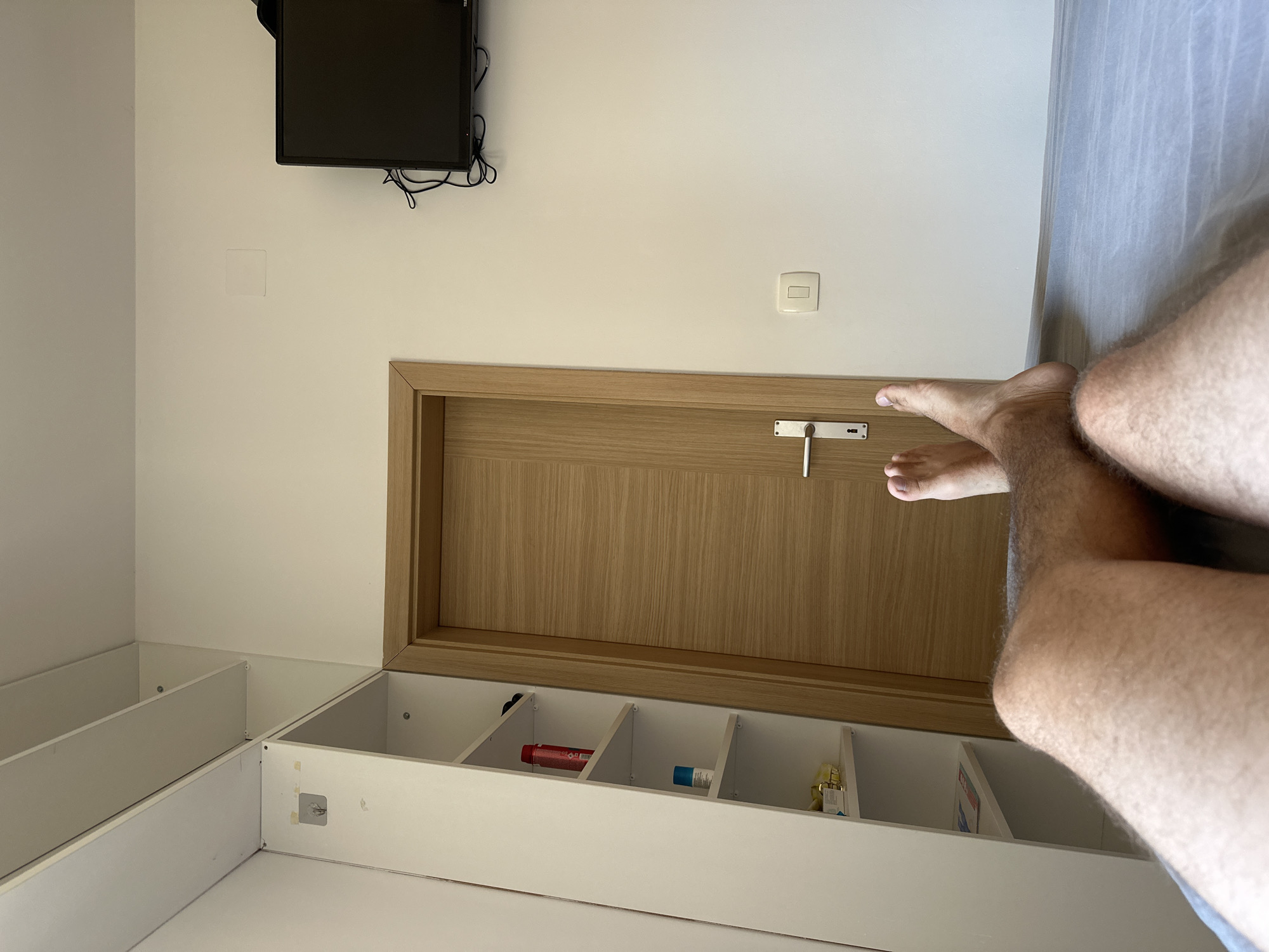 The image shows a person's legs and feet positioned near a door in a room. The room has white walls and a light wood door with a silver handle. To the left of the door is a white shelf unit with several compartments. Some items are visible in the compartments, including a red bottle, a blue tube, and a small yellow object. Above the door, a black television is mounted on the wall. A light switch is visible on the wall to the right of the door. The scene appears to be indoors, likely in a residential setting. The lighting suggests it is daytime. The overall impression is one of a simple, functional living space.