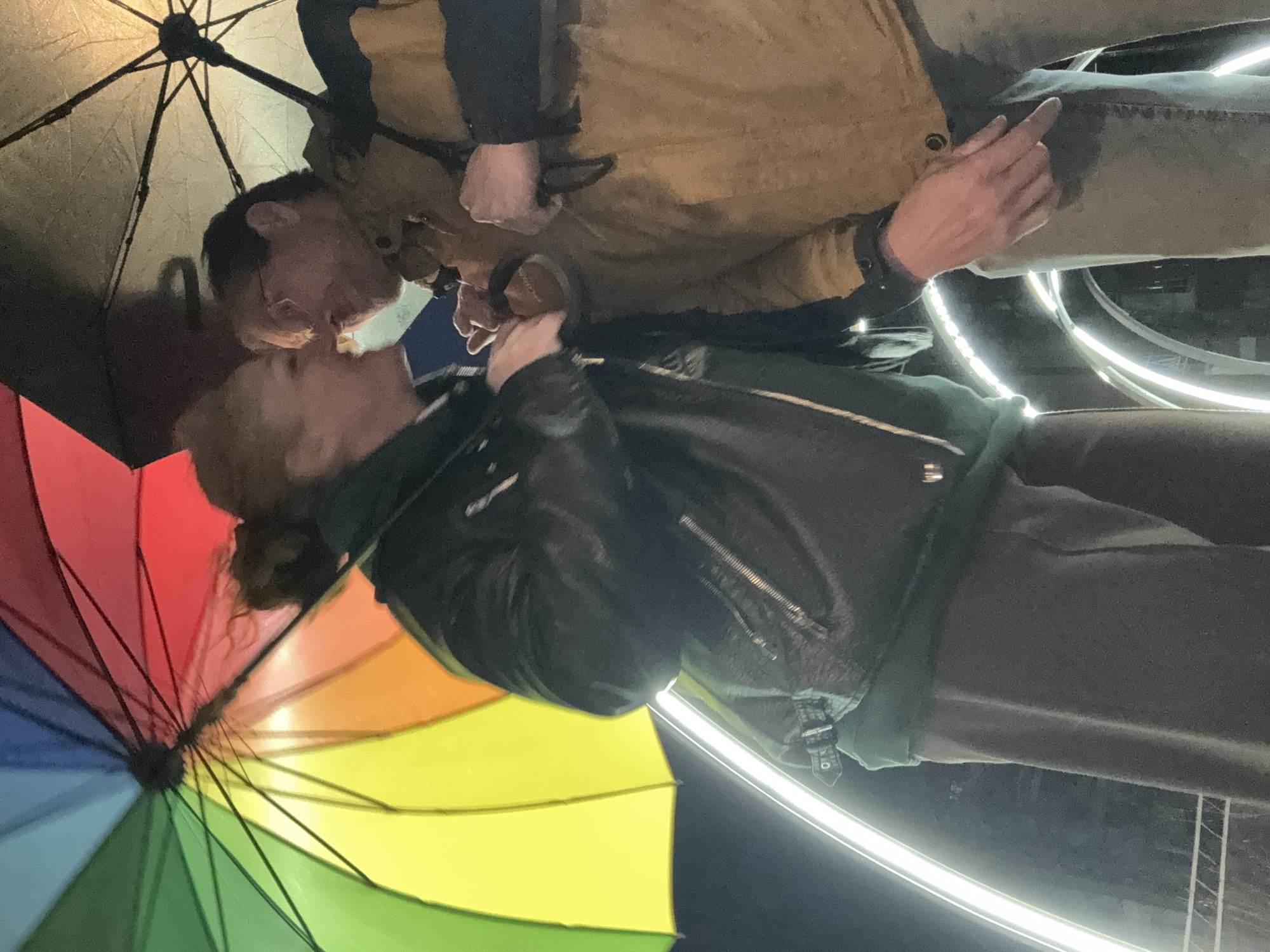A couple is kissing under a rainbow umbrella. The man is wearing glasses and a tan jacket. The woman is wearing a black leather jacket. They are likely standing in Chaikovsky, Russia, based on the prompt's request.