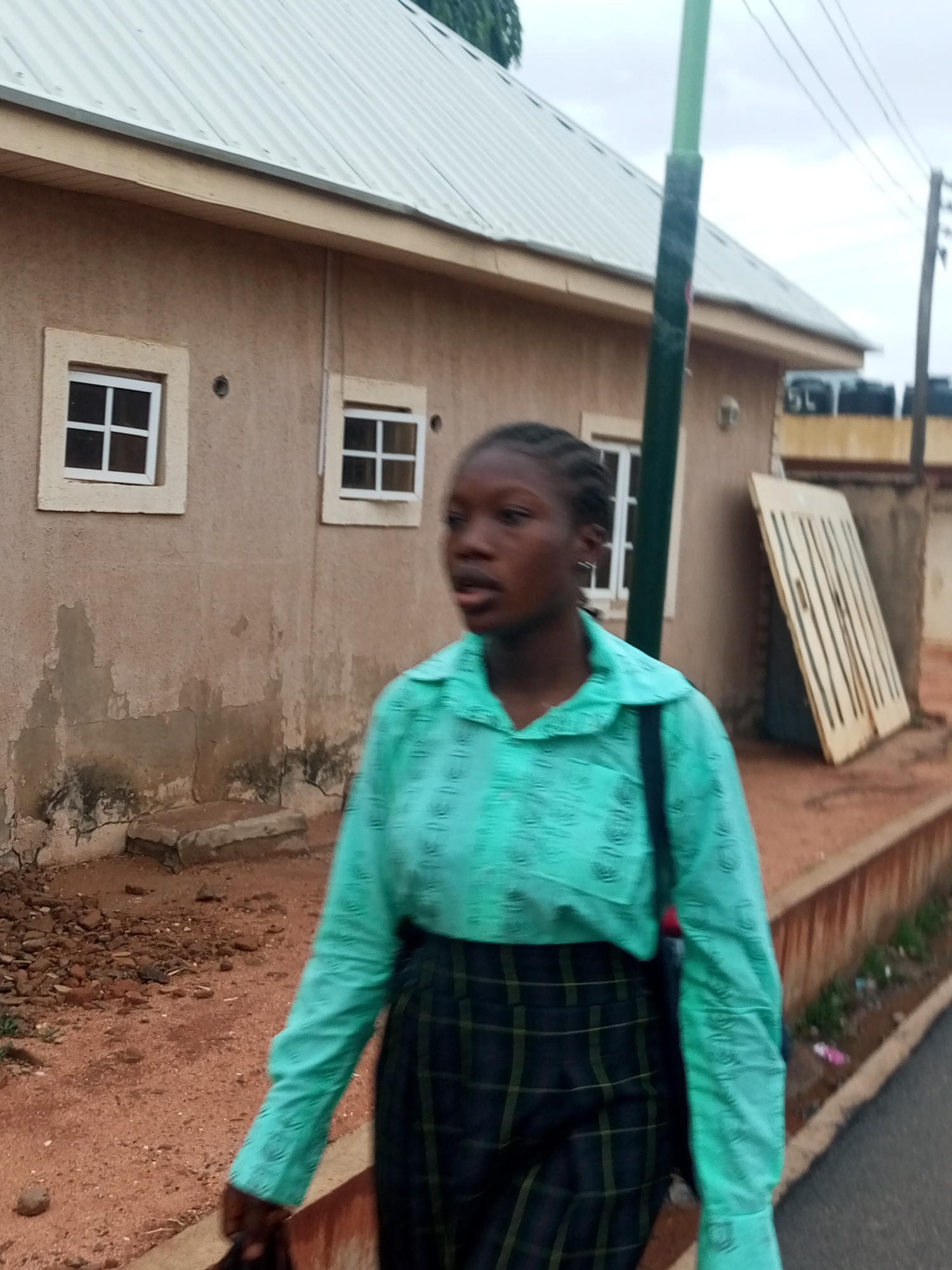 A young Black woman with braided hair, wearing a turquoise button-down shirt and plaid pants, walks on a street alongside a weathered building in Gombe, Nigeria. The building, which has a light-colored roof and small, square windows, shows signs of age and wear. Adjacent to the building is a green pole and a wooden structure. A curb separates the sidewalk from the street. Visible in the background are water tanks and power lines. The ground is a mix of dirt and pavement. The sky is overcast. The location is Gombe, Nigeria.
