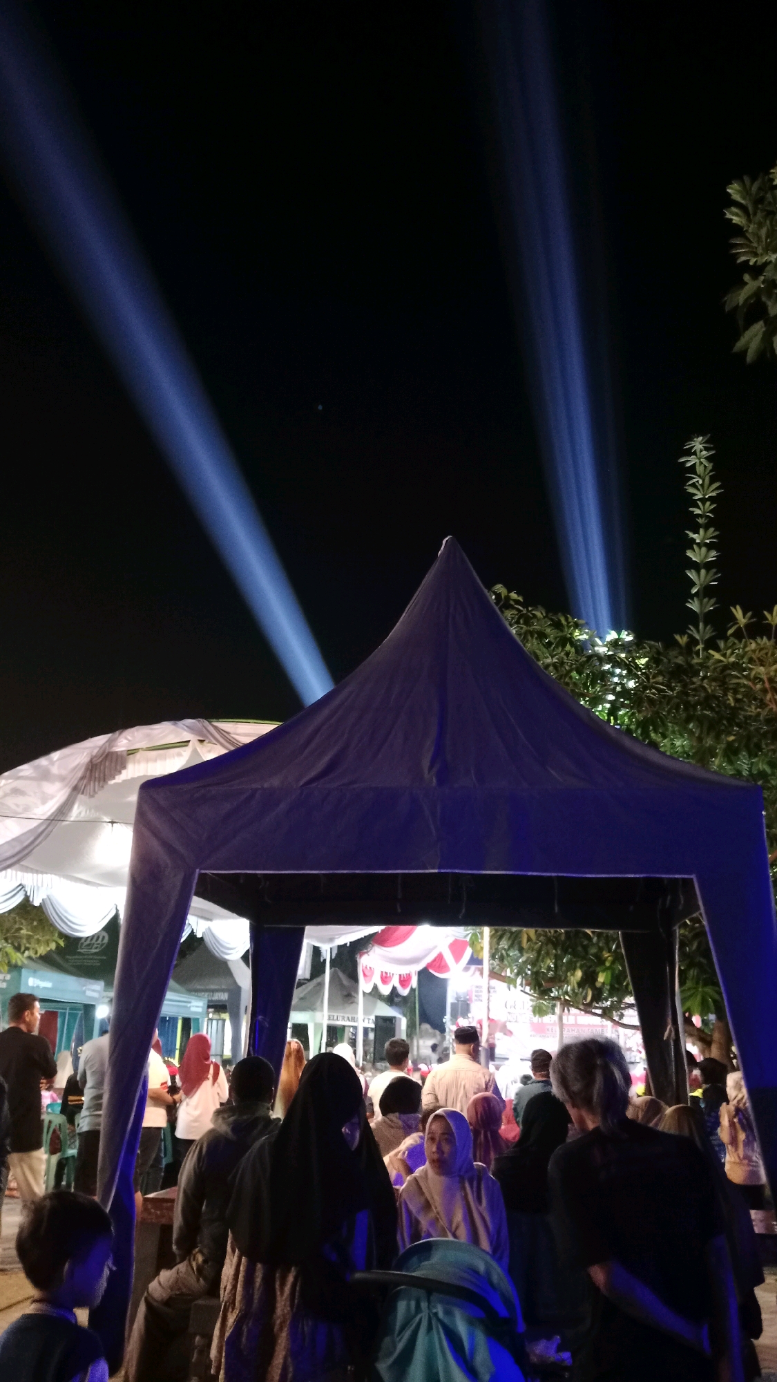 The image depicts a nighttime outdoor scene with a gathering of people, possibly a market or festival, in Tambran, Indonesia. In the foreground, a dark blue canopy tent structure is prominent, with a white tent to its left. Several individuals are visible, some standing and others seated. Many are wearing Islamic attire, including hijabs and headscarves. In the background, bright blue spotlights are projected into the night sky from an unseen source, creating dramatic beams of light that cut through the darkness. Trees are also visible to the right, with their leaves illuminated by the lights. Signs and structures are faintly discernible in the background, suggesting a commercial or event area. The overall atmosphere appears to be lively and illuminated by artificial light.