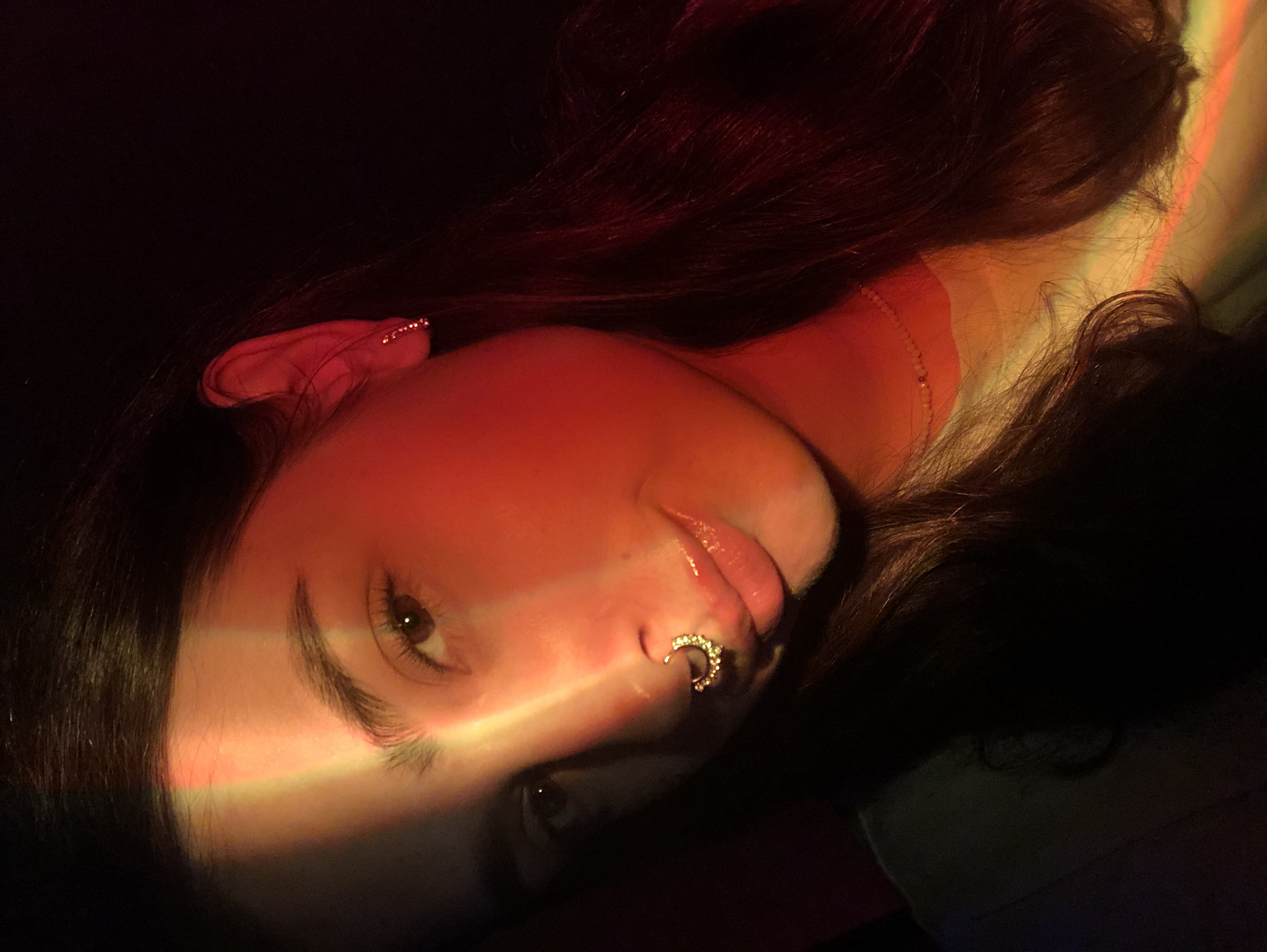 A young woman with long, dark hair is pictured in a close-up, selfie-style shot. She has a septum piercing adorned with small, glittering stones. Her left ear is visible, wearing a small, delicate earring. A beam of colorful light, featuring red and yellow hues, falls across her face, highlighting her nose and forehead. The background is dark, suggesting an indoor setting, possibly a dimly lit room or an environment with colored lighting. The image is taken from a slightly high angle, looking down at the woman. There is no discernible urban or geographical context that would specifically place it in Noviy, Russia.