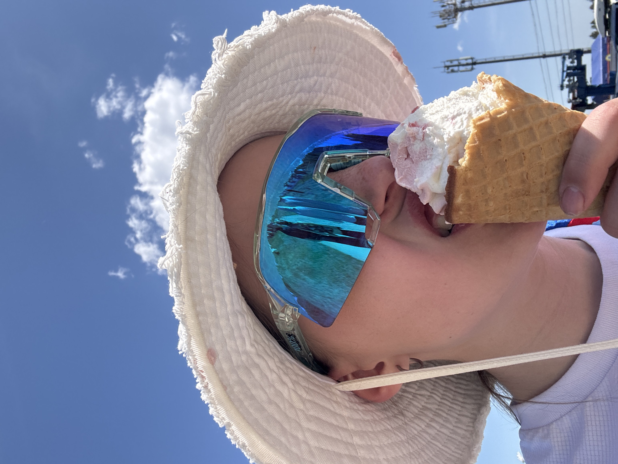 A person wearing a white hat and blue mirrored sports glasses is eating an ice cream cone. The cone is filled with white and pink ice cream. In the background, a blue sky with a few clouds is visible, along with a cellular tower structure. The location is likely Khvalynsk, Russia, based on the prompt.