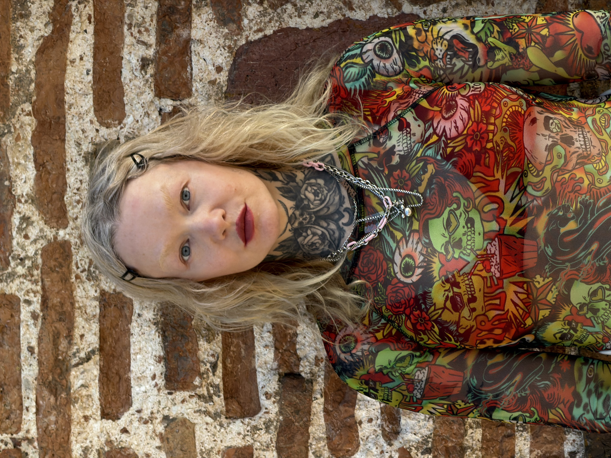 A person is lying on a surface of what appears to be brickwork. The person has pale skin, light blonde hair, and light eyebrows. Their neck is tattooed with dark ink patterns. They are wearing a mesh top or bodysuit with a colorful, patterned design that includes skulls, roses, and eyeballs. A chain necklace is visible around their neck. The background is a brick structure with variations in color and texture. The specific location in Hrodna, Belarus, cannot be determined from the image.