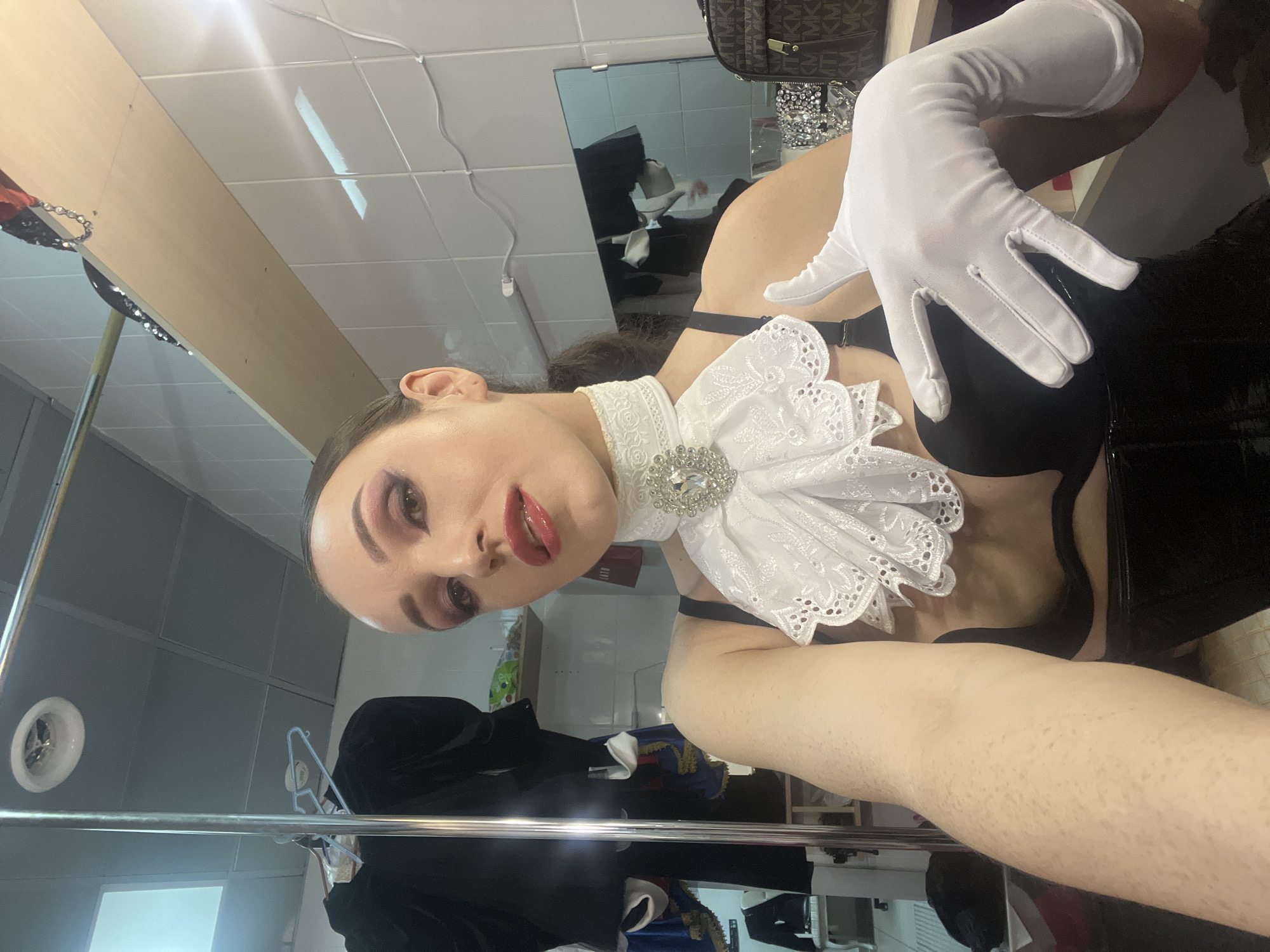 A person is taking a selfie in what appears to be a dressing room. The person has dark hair pulled back in a ponytail, and is wearing a white lace collar with a jeweled embellishment, a black bra, and white gloves. They are also wearing significant makeup with bright red lipstick and eye shadow. Behind them, clothing is hung on a rack, and there are shelves with various items, including a dark brown bag with a monogram pattern. The walls are tiled, and there is a mirror reflecting some of the room. The overall impression is one of preparation for a performance or event.