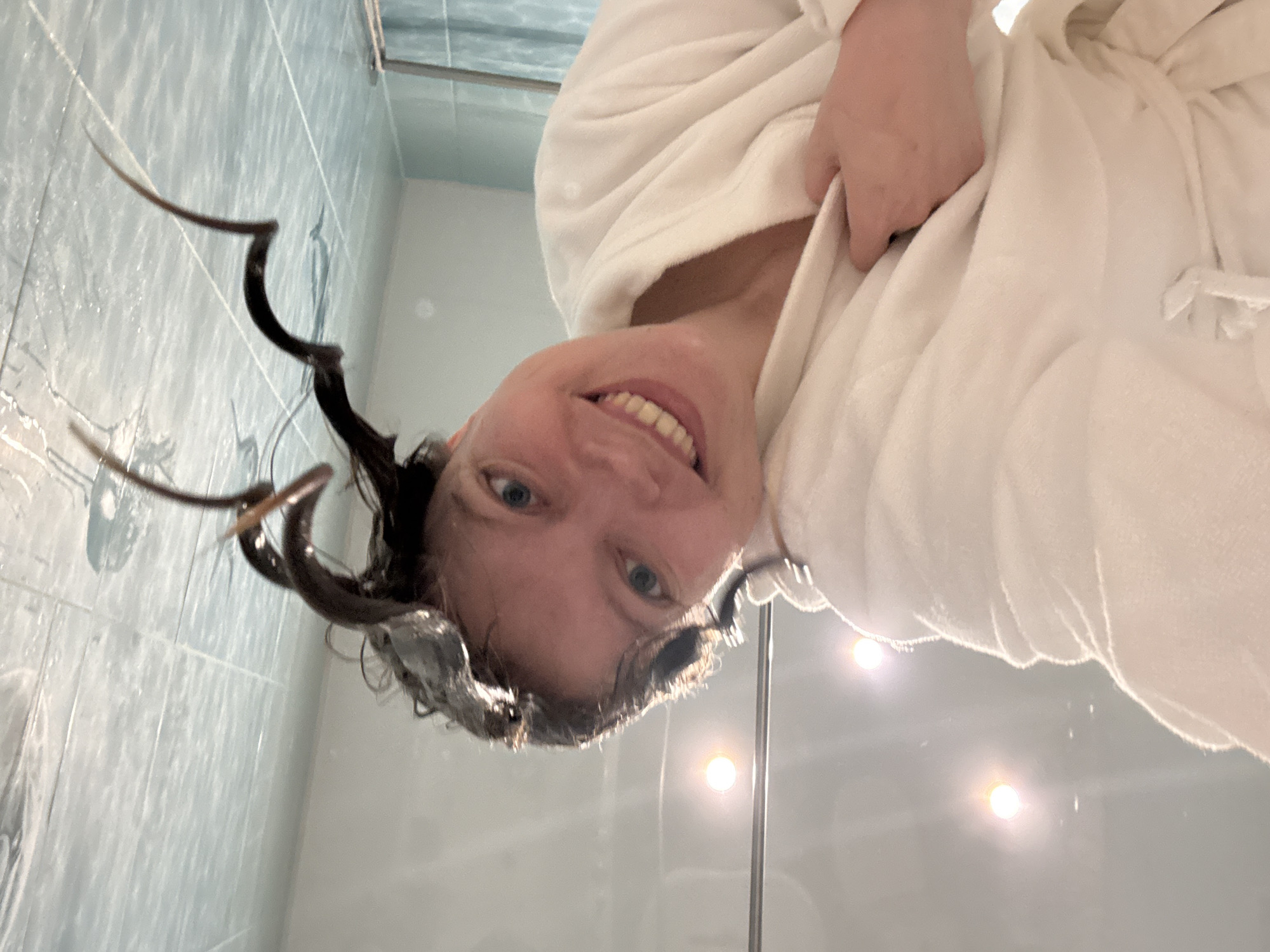 A woman with wet, dark, curly hair is wearing a white bathrobe. She is smiling at the camera. The background appears to be a bathroom with light blue tile and light reflecting off a shower door. The location is not discernible as Izhevsk, Russia, from visual information.