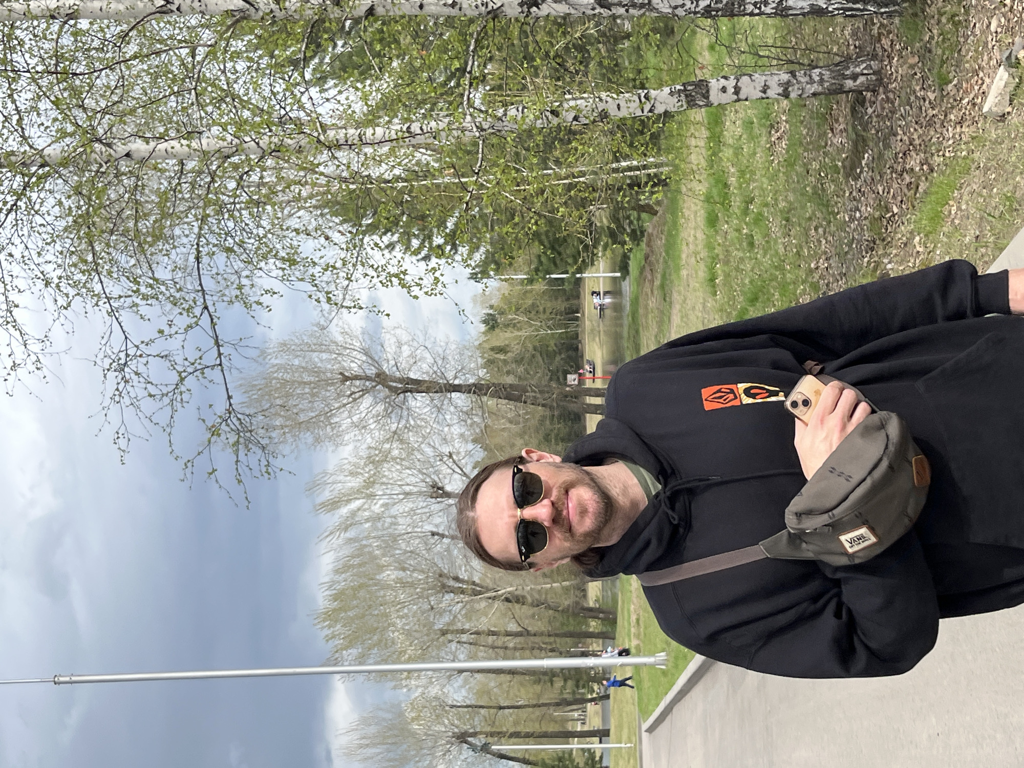 A man stands outside in Krasnoyarsk, Russia, wearing a black hooded sweatshirt with an orange and black logo, sunglasses, and a Vans fanny pack. He holds a smartphone in his hand. The backdrop consists of birch trees with light green leaves, a grassy area, a gray sky, and a gray concrete pathway.