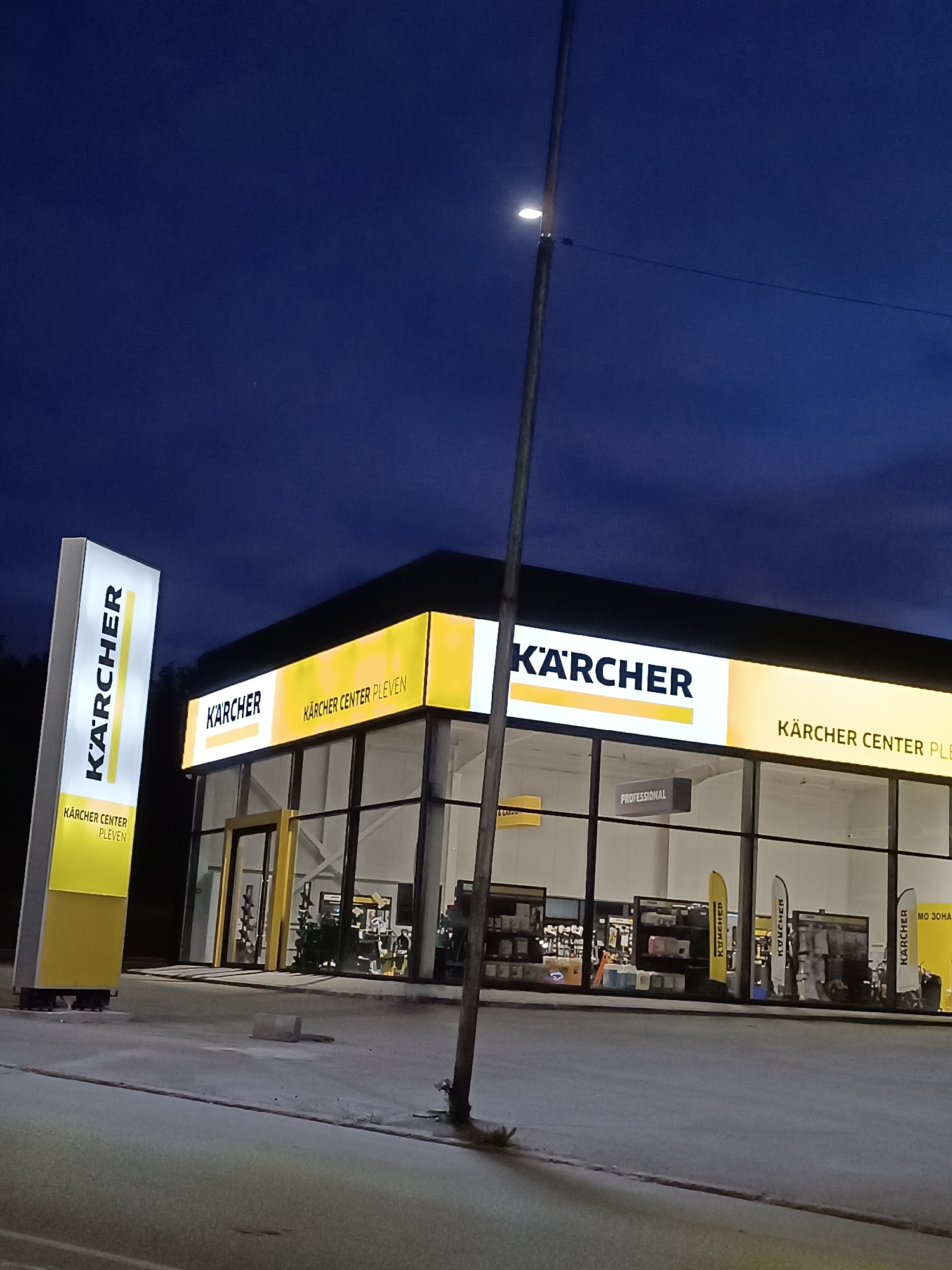 An exterior shot captures a Kärcher Center in Pleven, Bulgaria, at night or late dusk. The sky is a deep, dark blue, indicating the time of day. The modern commercial building is brightly illuminated with prominent yellow and white Kärcher branding.
The building features a large glass storefront, allowing visibility into the well-lit interior. The facade above the windows displays the Kärcher logo and text "KÄRCHER CENTER PLEVEN" in bold, glowing letters. Inside the store, Kärcher products are arranged on shelves, and several yellow and white feather flags bearing the Kärcher logo are visible. A hanging sign inside reads "PROFESSIONAL," and another partial sign to the right shows "МО ЗОНА."
To the left of the building stands a tall, illuminated pylon sign, also featuring the Kärcher logo vertically and "KÄRCHER CENTER PLEVEN" horizontally at its base. A street light pole, with its light on, stands in the foreground to the right, extending towards the center of the image and casting a subtle glow on the road. The immediate foreground consists of a grey paved surface, likely a road or parking area, with some subtle variations in texture. No people or vehicles are visible, giving the scene a quiet, almost serene ambiance.