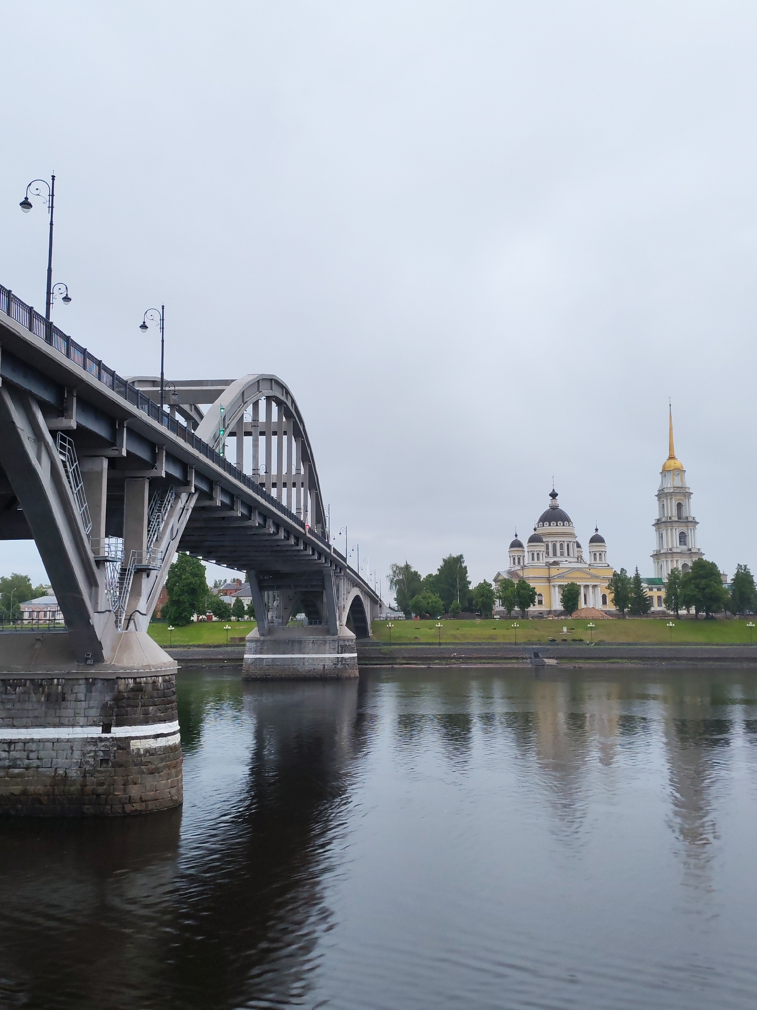 The image captures a cityscape of Bol'shie Mkhi, Russia. A concrete bridge dominates the foreground, its arches and support structures reflected in the dark water below. On the opposite bank, a yellow and white cathedral complex stands with a golden-topped spire adjacent to it. Trees are dispersed around the grounds. The sky is overcast. No people are visible in the scene.