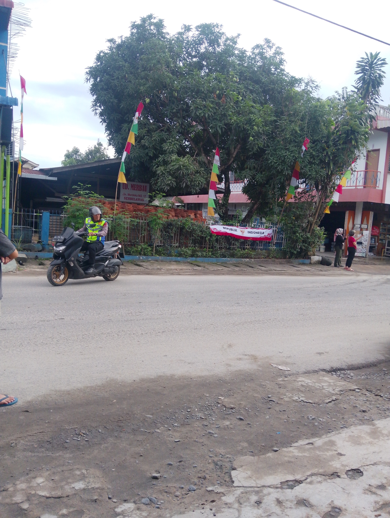 In Serbelawan, Indonesia, a man wearing a high-visibility vest and helmet rides a black motorcycle. He is on a paved road, which shows signs of wear and tear. Behind him, a building with a pink facade and balconies stands beside a large tree adorned with colorful triangular flags. A fence separates the building from the road, and a banner reading "REPUBLIK INDONESIA" is visible. Several people are seen near the building, including two women and a person whose arm is visible in the foreground. The sky is overcast, and the surrounding vegetation is lush.