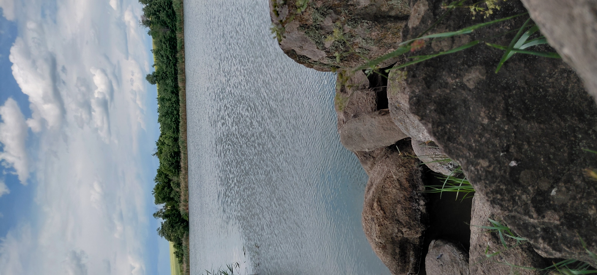 A serene natural landscape unfolds in this vertically oriented image, capturing a tranquil body of water bordered by lush greenery and rocky formations. The water, rippling gently, reflects the soft, diffused light of an overcast day. A dense line of trees and vegetation runs along the far bank, creating a natural barrier between the water and a distant, unseen landscape. To the right, large, weathered rocks of varying sizes dominate the foreground, their rough textures and earthy tones contrasting with the smooth expanse of the water. Patches of moss and lichen cling to the rocks, suggesting a humid environment and the passage of time. Wisps of green grass sprout from crevices in the rocks and along the water's edge, adding a touch of life to the scene.
The sky above is a canvas of soft blues and whites, with clouds drifting lazily, indicating a peaceful, perhaps slightly cool, weather condition. The overall atmosphere is one of stillness and natural beauty, inviting contemplation. The image is devoid of any human presence or man-made objects, emphasizing the untouched quality of the natural setting. The location cues, such as the specific type of vegetation and the rocky terrain, along with the provided context of Novomykolaivka, Ukraine, suggest a rural or semi-natural area within that region. There is no visible text in the image.