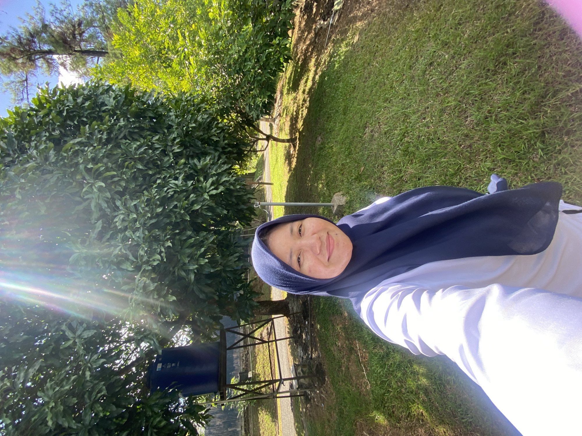 A selfie features a woman wearing a navy blue hijab and a white shirt. She is standing outdoors in a grassy area with trees in the background. Sunlight is streaming through the trees, creating lens flare. To the left of the woman is a large blue water tank on a metal stand. The location appears to be a park or a residential area with lush vegetation. The scene is well-lit, suggesting it is daytime. The woman is smiling at the camera. The environment suggests a tropical or subtropical climate. Banjarbaru, Indonesia is known for its green spaces and agricultural areas, consistent with this image.