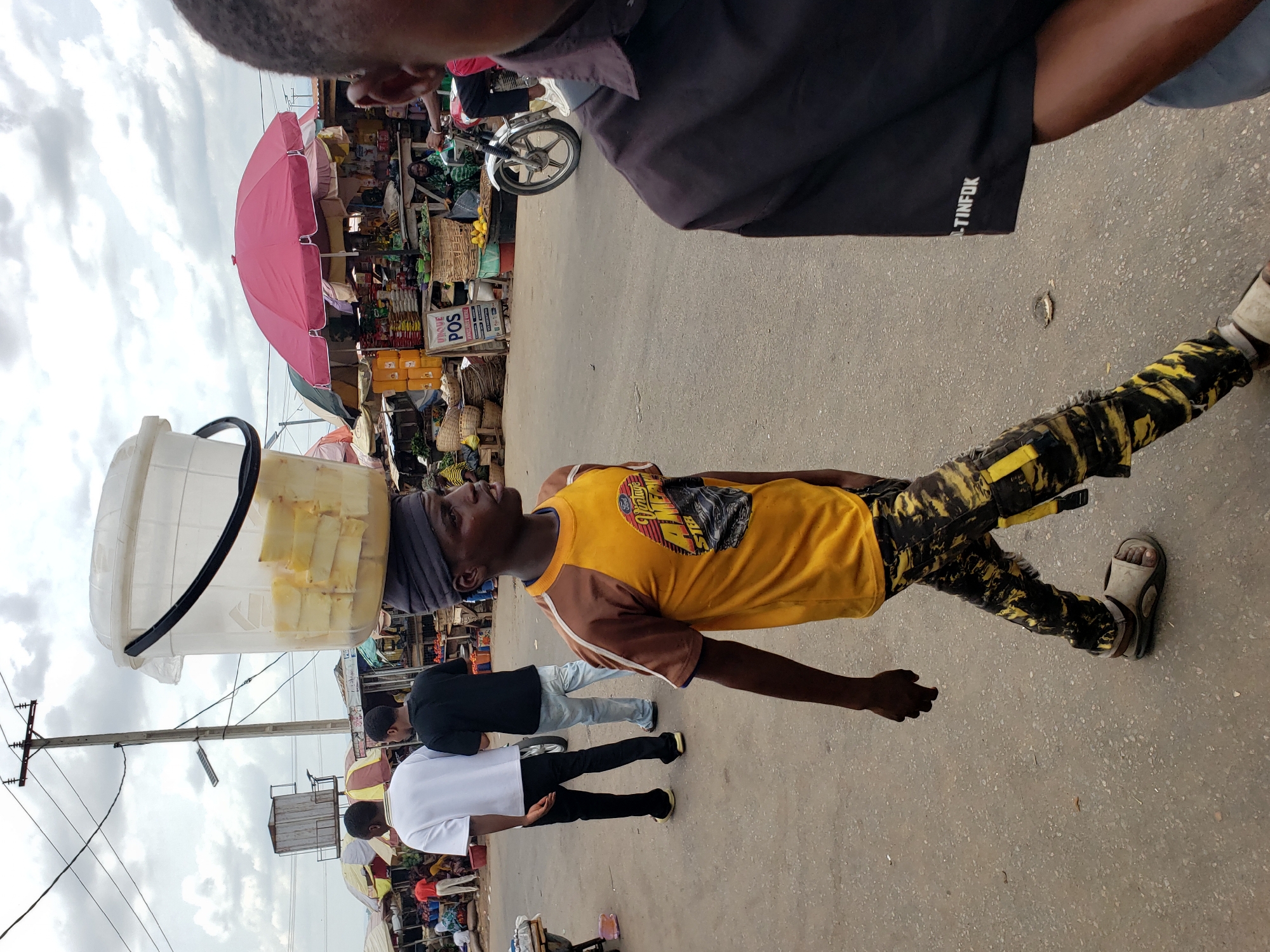 The image captures a bustling street scene in what appears to be a market area in Lokoja, Nigeria, under an overcast sky. The primary subject in the mid-ground is a young man walking towards the right, expertly balancing a large, clear plastic bucket filled with cut yellow fruit (likely pineapple) on his head. He wears a yellow and brown short-sleeved t-shirt, distinctive yellow and black patterned trousers, and light-colored sandals. A dark cloth is wrapped around his head, presumably to cushion and stabilize the load. He looks upwards and slightly to his right, maintaining his focus.
Behind this fruit seller, further to the left, two other men are walking. One wears a black t-shirt and light blue jeans, while the other is dressed in a white t-shirt and dark trousers. In the top right foreground, the upper back and arm of another person are visible, wearing a dark shirt that has "TINFOK" printed on its sleeve.
The background is rich with details of a lively market. A prominent red market umbrella provides shade over a stall. Numerous market stalls are visible, displaying various goods including baskets, packaged items, and other produce like yellow fruits in a basket. A sign bearing the text "UNIQUE POS" is clearly visible on one of the stalls. A motorcycle is partially seen behind the market setup. Further to the left, a utility pole with electrical wires and what appears to be a water storage tank on a stand contribute to the urban landscape. The ground beneath the people is a paved road. The scene portrays a typical daytime market environment, highlighting local commerce and daily movement.