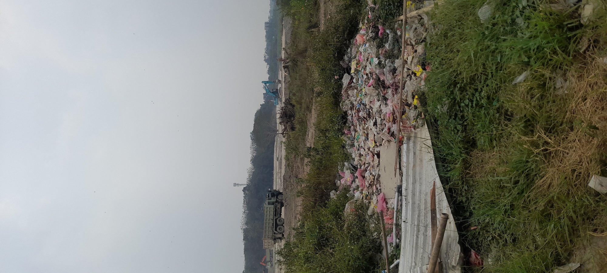 The image depicts an outdoor scene with a large pile of garbage in the foreground, primarily consisting of plastic bags and debris, interspersed with some vegetation. In the middle ground, a construction site is visible with a large truck and an excavator. The background features a treeline and a hazy sky, with a cell tower in the distance. The ground appears to be dry and dusty, consistent with a construction area. The overall impression is one of an undeveloped or under-construction area with significant litter.