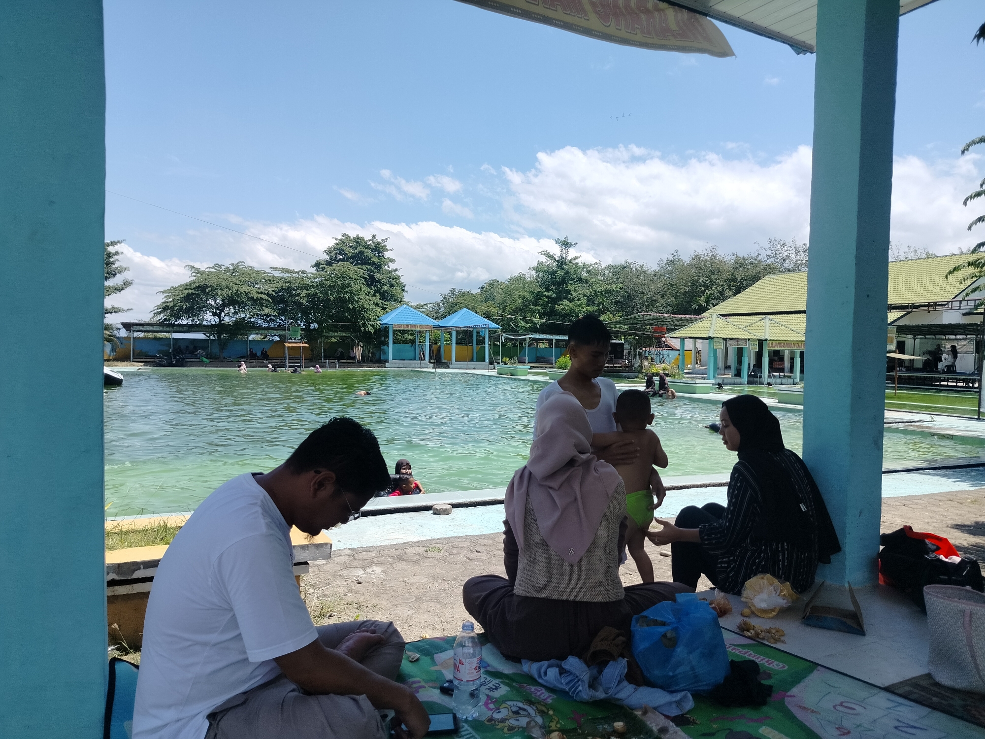 This image captures a sunny day at a swimming pool complex in Tegarmiko, Indonesia. In the foreground, a man in a white t-shirt and light-colored pants is seated on the ground, looking down at his phone. To his right, a woman wearing a light pink hijab and patterned shirt is also seated, facing away from the camera. Beside her, a child in a green diaper is being held by a person whose face is obscured. Another woman in a black patterned outfit is seated to the right of the child, looking towards the pool. A blue plastic bag, a water bottle, and some food items are scattered on a patterned mat in front of the seated individuals.
The swimming pool, filled with green water, is visible in the midground, with several people swimming and wading. The pool area is lined with a paved walkway and bordered by trees and buildings. In the background, there are structures with blue and green roofs, typical of recreational facilities. The sky is bright blue with scattered white clouds. The overall scene suggests a relaxed, family-oriented outing at an outdoor pool.