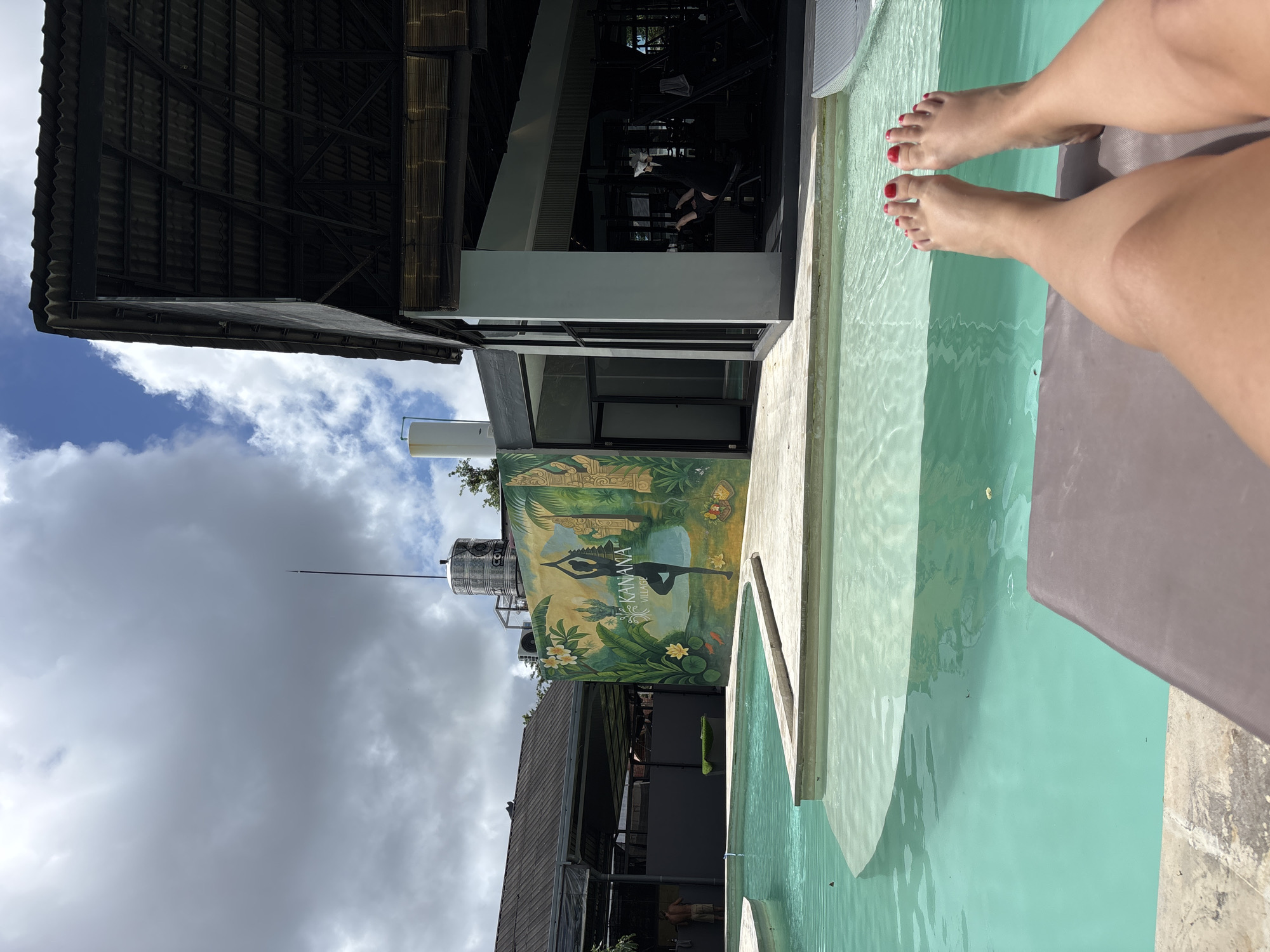 The image depicts a view overlooking a swimming pool at a facility identified as “Kanana Village” in Kuta Selatan, Indonesia, as suggested by the signage on a nearby structure. The vantage point is from a person reclining poolside, with their bare feet painted with red nail polish visible in the foreground. The pool water is turquoise in color. Adjacent to the pool is a building that features a mural depicting a figure in a yoga pose amidst lush tropical vegetation. Above the mural, a cylindrical tank is visible with a thin antenna extending upwards. The architecture of the buildings suggests a modern design with open structures. The sky overhead is partly cloudy.