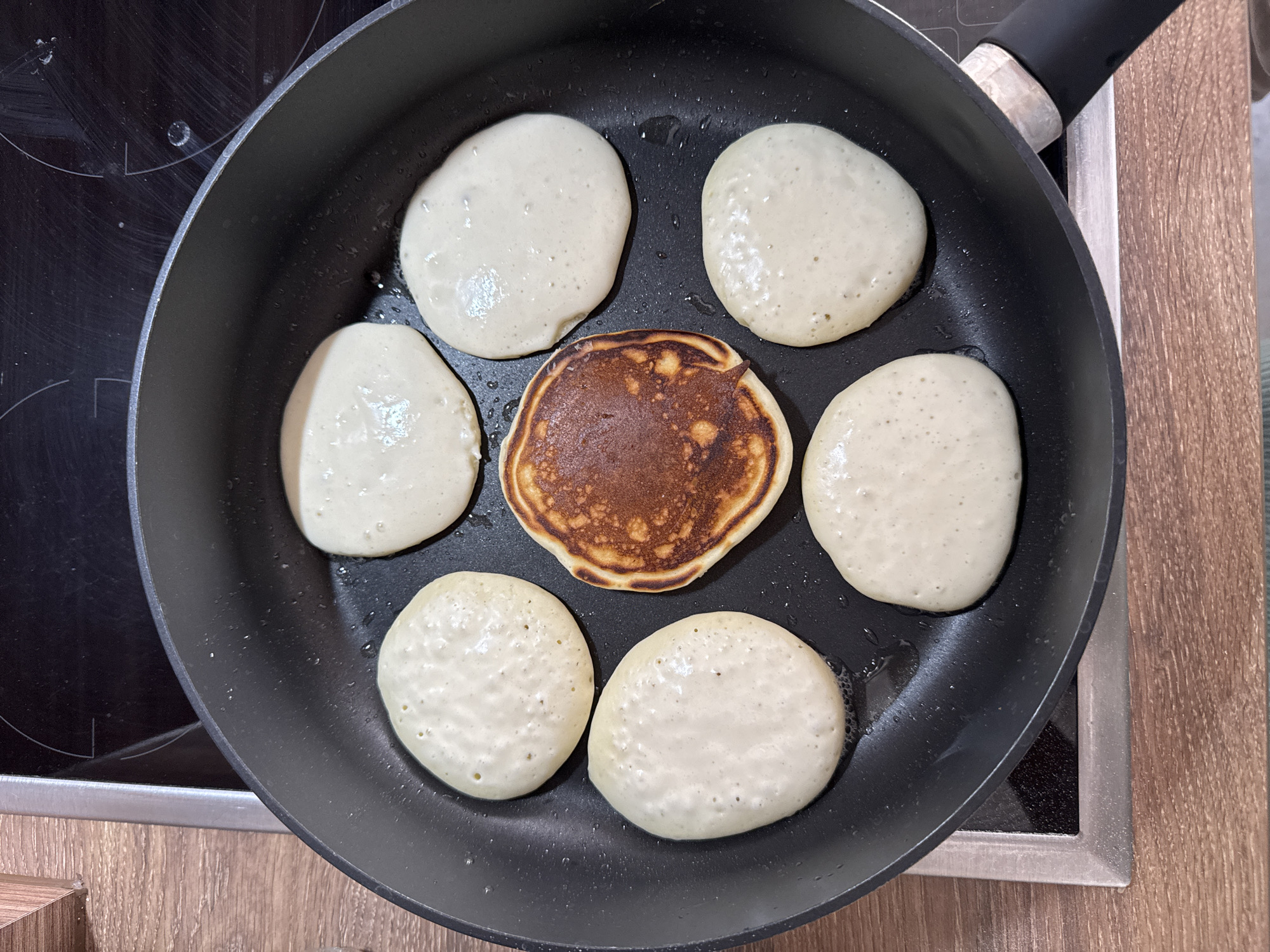 A top-down shot shows seven pancakes in a circular arrangement within a black frying pan. Six of the pancakes are uncooked, with a pale, slightly bubbly batter. One pancake in the center is golden brown and appears to be cooked. The pan is resting on a black glass stovetop, and a wooden countertop is visible on the left. The overall scene suggests food preparation.