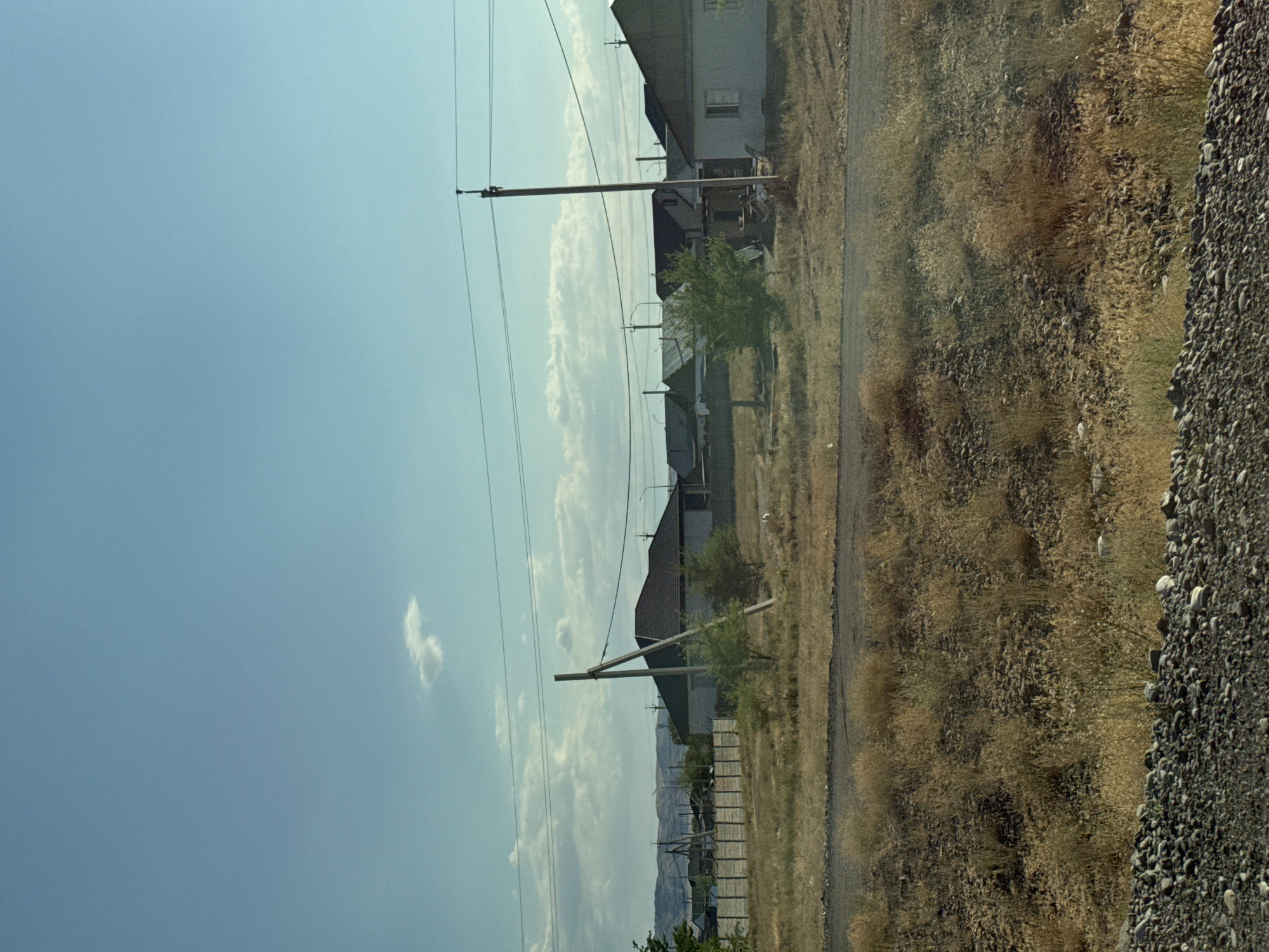 This is a photograph of a residential area in the city of Usharal, Kazakhstan, on a clear day with a pale blue sky and scattered white clouds. The foreground is dominated by dry, golden-brown grass and gravel. A dirt road or path runs horizontally across the middle ground. Beyond the road, a line of houses with dark-colored roofs and light-colored walls are visible. A concrete fence runs along the left side of the frame, separating the dry vegetation from the houses. Several utility poles with power lines are present throughout the scene, including a prominent one with diagonal bracing in the center right. Trees and bushes are interspersed among the houses and along the fence line. The background features rolling hills under a hazy sky.