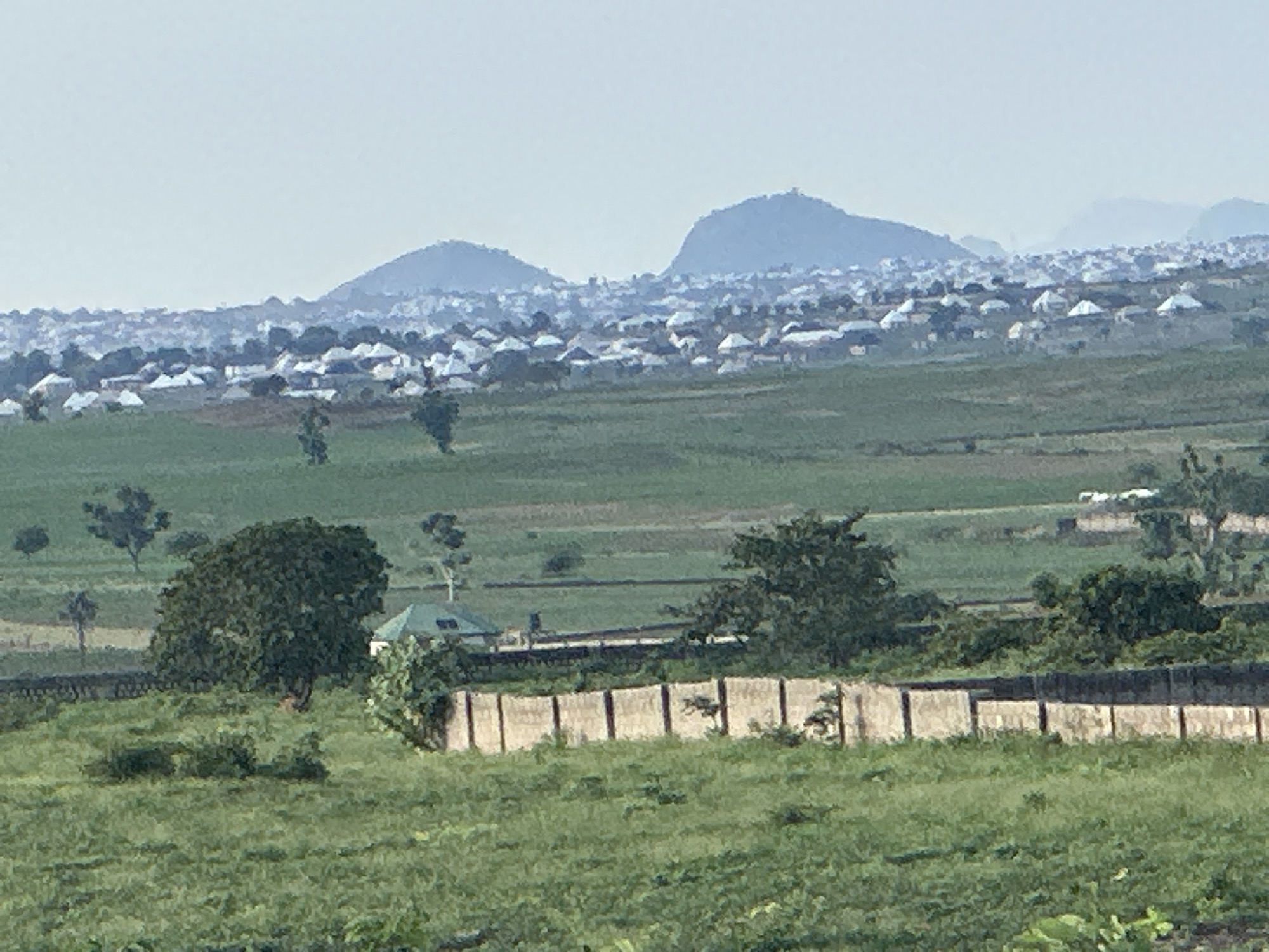 The image depicts a vast, hazy landscape dominated by rolling green hills and a distant settlement. In the foreground, a fence made of concrete posts and panels runs horizontally across the frame. Beyond the fence, the terrain is covered in lush green grass, with scattered trees. A prominent, large tree with dense foliage stands to the left of the frame, near a small green-roofed building.
Further in the background, a sprawling urban area is visible, characterized by a dense collection of houses with white roofs, interspersed with trees. The houses appear to be low-rise structures. Behind the settlement, a range of majestic blue-hued mountains rises, their peaks softened by the atmospheric haze. The sky is a pale, muted blue, suggesting either early morning or late afternoon light. The overall impression is one of a rural or semi-rural setting at the edge of a populated area, likely Gidan Waziri, Nigeria, given the context provided. No people are visible in the image.