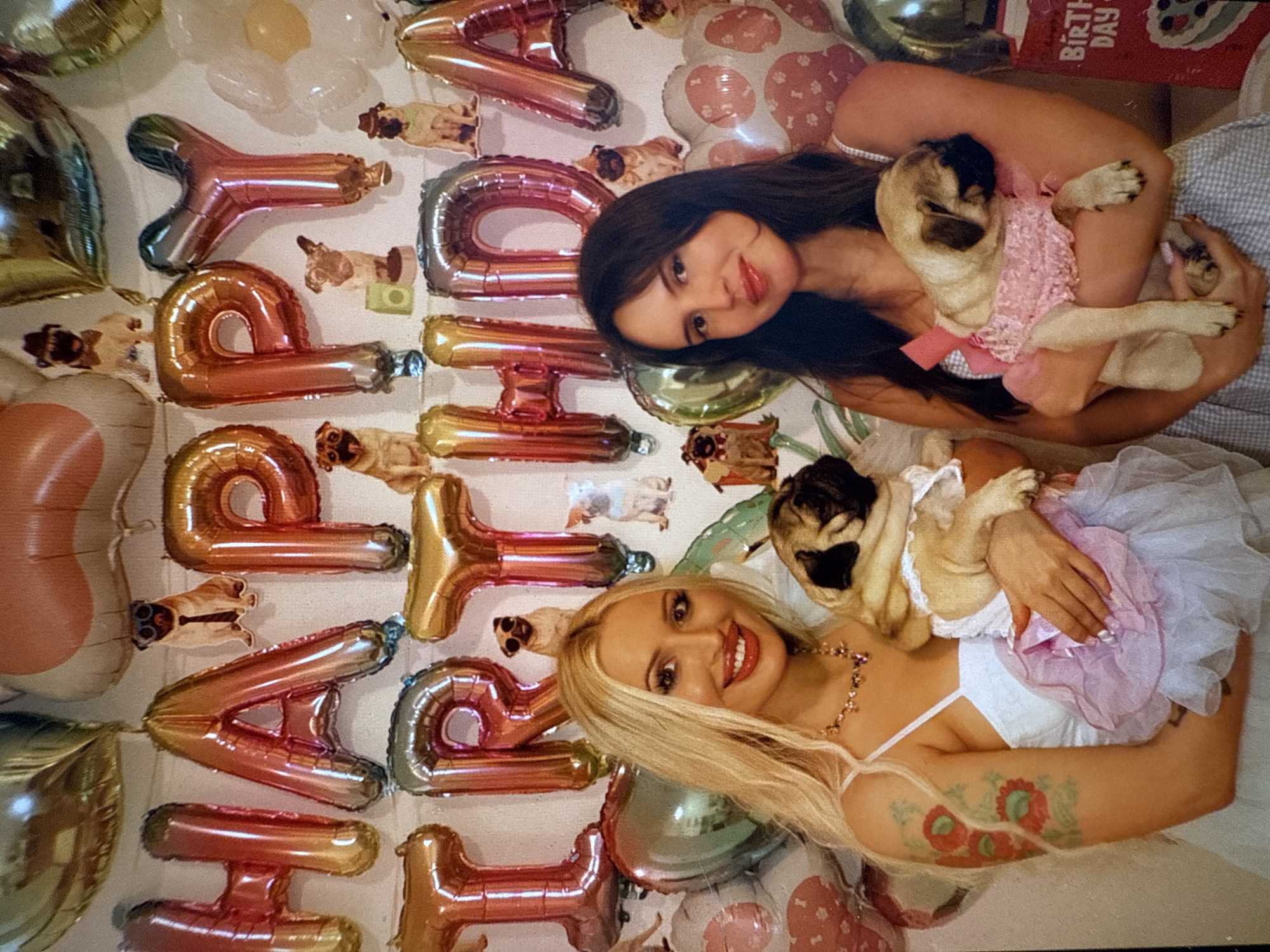 Two women and their pug dogs are captured in an indoor setting, celebrating what appears to be a birthday party. The image is oriented diagonally, with the two women positioned side-by-side, each holding a pug.
The woman on the left has long dark hair and is wearing a light-colored top. She holds a fawn-colored pug that is dressed in a sparkly pink dress with a bow. This pug is looking upwards and appears relaxed in her arms.
The woman on the right has long blonde hair, styled straight, and is wearing a white bralette-style top. She has a prominent floral tattoo on her upper left arm. She holds a fawn-colored pug wearing a white top and a pink and white tutu-like dress. This pug is also looking upwards and seems comfortable. Both women are smiling warmly at the camera.
The background is adorned with numerous foil balloons spelling out "HAPPY BIRTHDAY" in rose gold and iridescent colors. Scattered among the balloons are several small cut-out images of pugs in various poses, reinforcing the dog-centric theme of the celebration. There are also other decorative balloons, including round ones and some with paw print or bone designs. In the upper right corner, a red box or card with "BIRTHDAY" printed on it is visible. The overall atmosphere is joyful and celebratory, centered around the beloved pets.