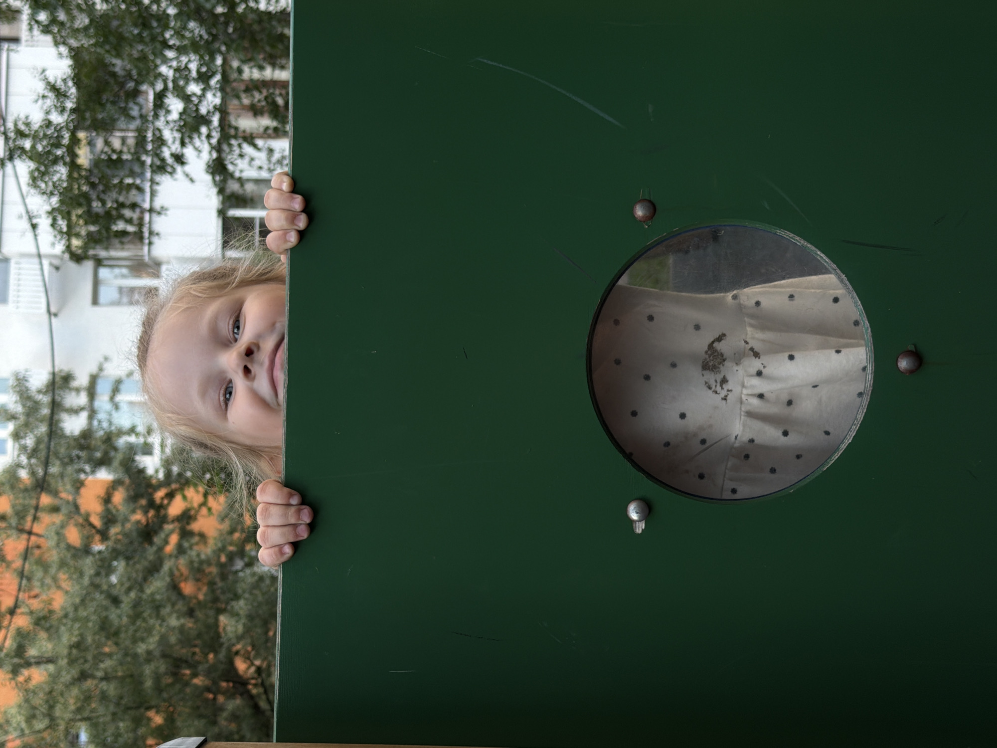 A young girl with blonde hair peeks over a green fence on a playground. Her hands grip the top edge of the fence. She is looking directly at the camera with a slight smile. A circular window is visible in the fence below her, through which a portion of her white dress with dark polka dots can be seen. The background consists of trees and buildings, indicative of an urban environment in Moscow, Russia.