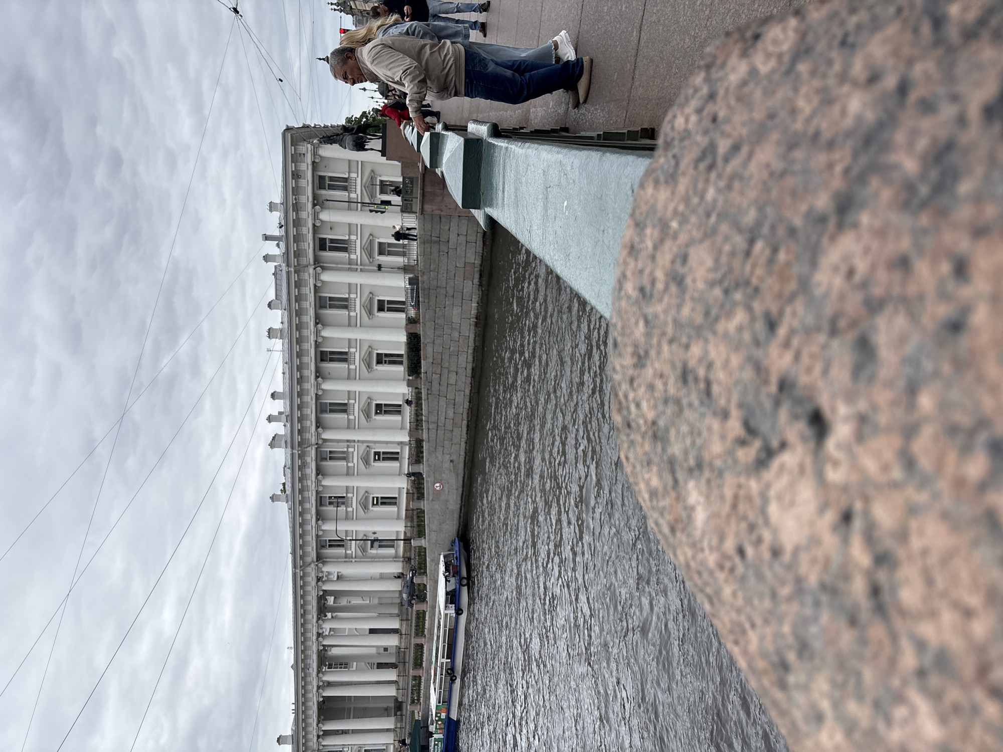 The image, taken from a low angle on a bridge, shows a man leaning over the bridge railing, seemingly looking at the water. The bridge railing is green and attached to a large, granite structure. In the background, a large, white building with rows of columns faces the river. A boat is seen on the water, traveling along the building's edge. The sky is overcast and gray, suggesting cloudy conditions. The location is Saint Petersburg, Russia.