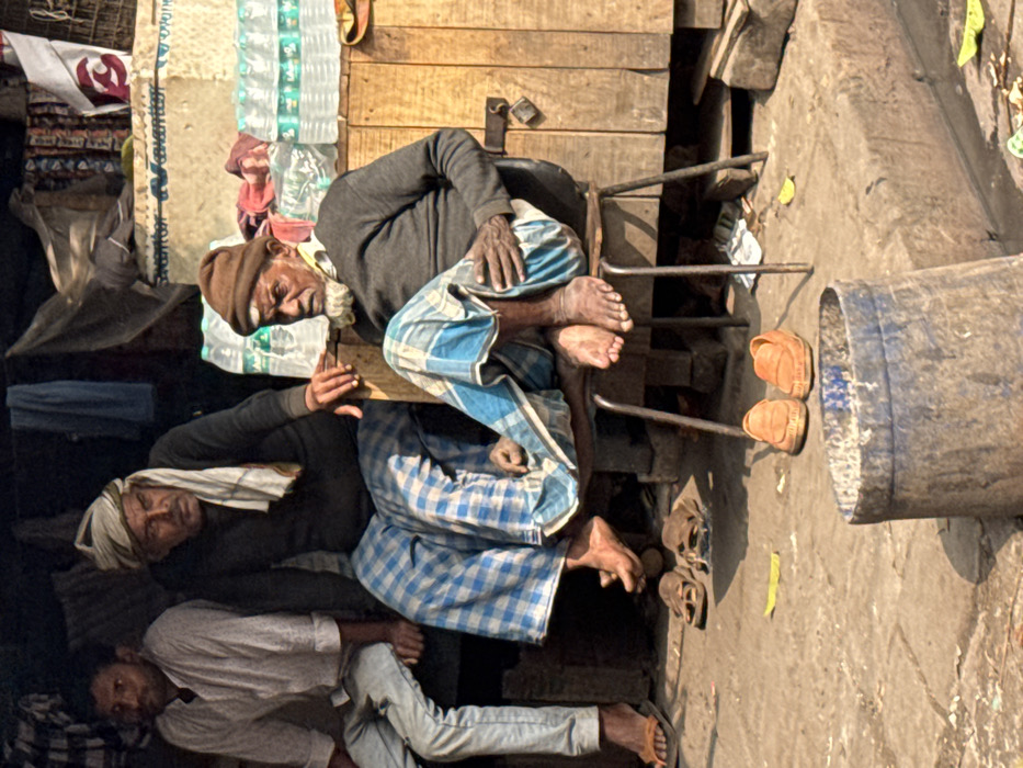 Three men lay together in close quarters. One man, clad in a blue and white patterned cloth, reclines on a makeshift platform. His brown-toned skin is wrinkled with age. A second man beneath him wears a gray shirt and a white cloth wrapped around his head. The third man wears a white shirt and matching pants. A pair of shoes sits on the ground near a bucket, positioned next to the men.
