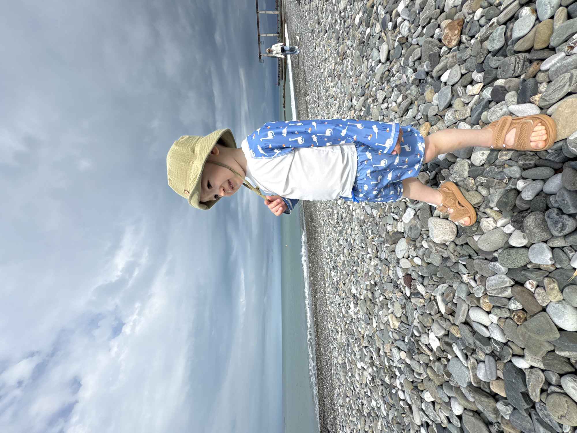 A young child stands on a pebble beach, possibly in Sochi, Russia. The child is wearing a light-colored hat, a blue and white long-sleeved shirt with dinosaur or animal print, and brown sandals. The beach consists of numerous grey and white pebbles. In the background, the sea meets a cloudy sky. Further back, a pier or structure can be seen with indistinct figures on it.