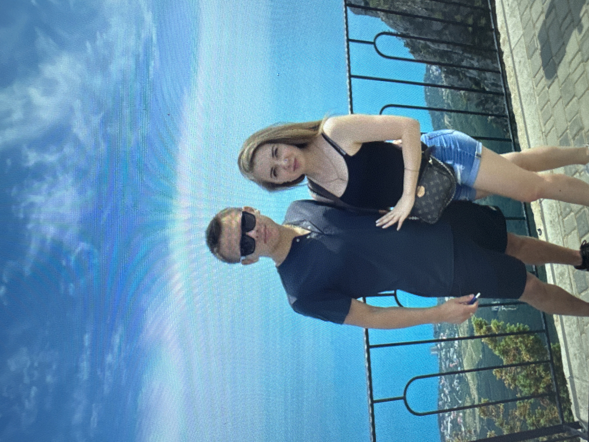 A young couple is posing for a photo on an elevated walkway overlooking a town and a body of water under a bright, blue sky with scattered clouds. The man on the left wears a dark blue t-shirt and black shorts, with black sunglasses and dark hair. The woman on the right wears a black tank top and denim shorts, with long blonde hair and a small crossbody bag. A black metal railing is in the foreground, separating them from the view. The walkway is paved with light-colored bricks. The background shows a coastal town with buildings nestled on hillsides, lush green trees, and a vast expanse of bright blue ocean.