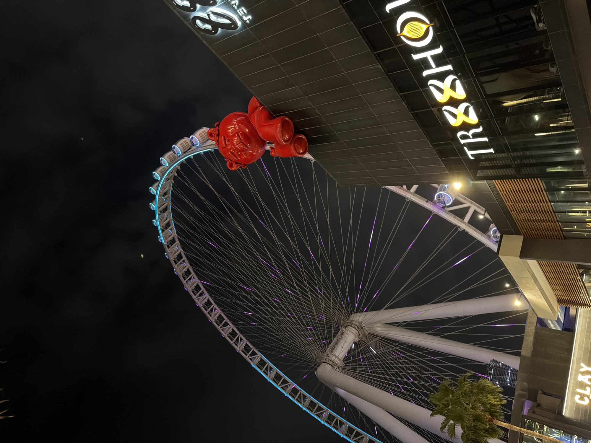 The media file presents a vibrant nighttime scene captured with a tilted perspective, looking upwards at a large Ferris wheel and an adjacent modern building. The dark sky fills much of the upper left portion of the image.
The dominant feature is a massive Ferris wheel, partially visible across the left and bottom of the frame. Its outer structure is outlined with bright blue lights, while numerous spokes radiate from its center, illuminated with a soft purple glow, creating an intricate, web-like pattern. Several illuminated passenger cabins are visible along its rim.
A contemporary building, with dark, tiled surfaces and wooden slat accents, occupies the top and right side of the image. A striking red sculpture is affixed to this building's facade, appearing to "climb" or hang. This large, stylized figure resembles a cartoon character or mascot, featuring a prominent head with visible facial features and large, rounded appendages.
The building displays several illuminated signs. A prominent neon sign at the top reads "TR88HOUSE" in white and yellow lettering, accompanied by a stylized leaf or flame logo. Further down on the right, another illuminated sign clearly spells "CLAY". Several palm trees are visible near the base of the building on the right.
The scene depicts a bustling urban environment at night, illuminated by artificial lights, indicative of an entertainment or leisure complex. The notable elements and architecture are consistent with locations like Ain Dubai in Dubai, United Arab Emirates.