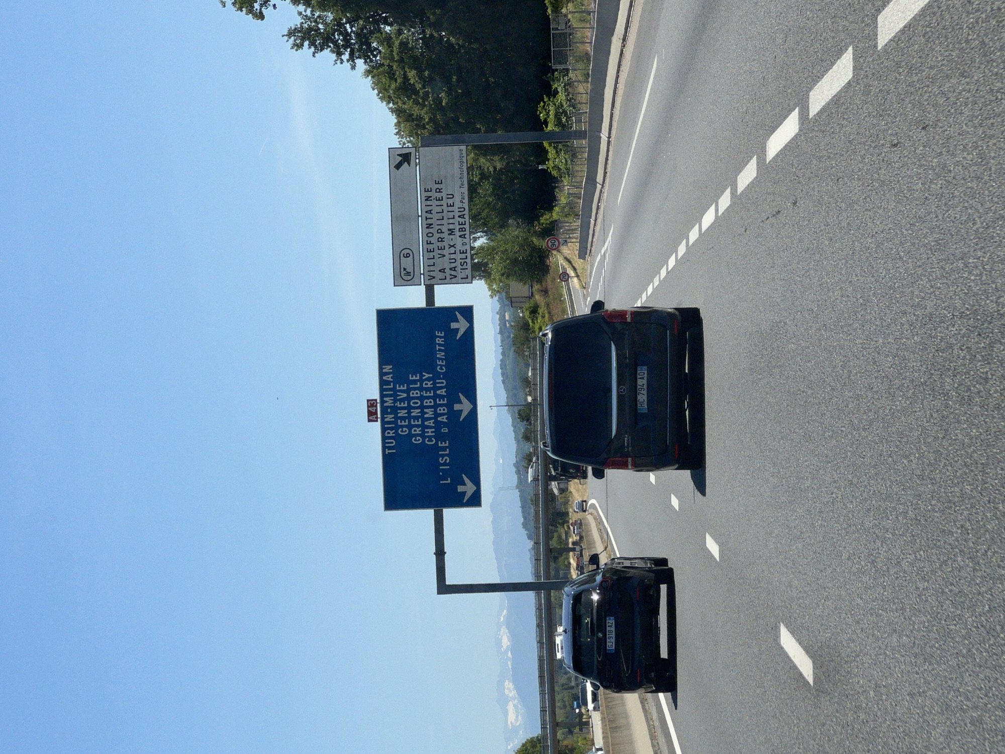 The image shows a highway scene with a bright blue sky. In the foreground, two dark-colored SUVs are driving on the road. The SUV on the left has a license plate that reads "GJ 918 AZ". The SUV on the right has a license plate that reads "HC 794 AQ". In the background, there are tall green trees and distant mountains. Overhead, there are two large highway signs. The sign on the left, marked with "A 43", lists destinations: "TURIN-MILAN", "GENEVE", "GRENOBLE", "CHAMBERY", and "L'ISLE D'ABEAU-CENTRE". The sign on the right indicates "N 6" and lists destinations: "VILLEFONTAINE", "LA VERPILLIERE", "VAULX-MILIEU", and "L'ISLE D'ABEAU-Parc Technologique". There are also speed limit signs visible on the side of the road. The overall scene depicts a typical highway environment with directional signage.