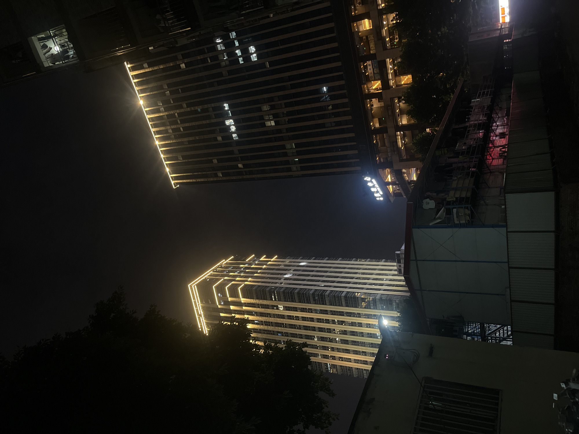 The image captures a nighttime view of urban buildings in Yuzhong District, China. In the foreground, a low-rise structure with a barred window and visible electrical conduits is partially illuminated. To its right, another temporary-looking structure with a corrugated metal fence in front of it displays glowing red lights from within.
Dominating the mid-ground and background are two tall, modern buildings. The building on the left is a skyscraper with vertical lighting that outlines its edges, giving it a glowing appearance against the dark sky. The building on the right is also a skyscraper, featuring a facade of vertical dark lines and illuminated windows. The upper portion of this building is also outlined with bright yellow lighting.
Below the right skyscraper, a multi-story building with illuminated floors, likely commercial or office space, is visible. A sign with Chinese characters and English text, "Yuzhong CBD," is mounted on the facade of this lower building. Trees are scattered between the foreground structures and the skyscrapers, their dark silhouettes contrasting with the artificial lights. The overall scene is a juxtaposition of older, simpler structures against the backdrop of modern, brightly lit high-rise architecture, characteristic of an urban night environment.