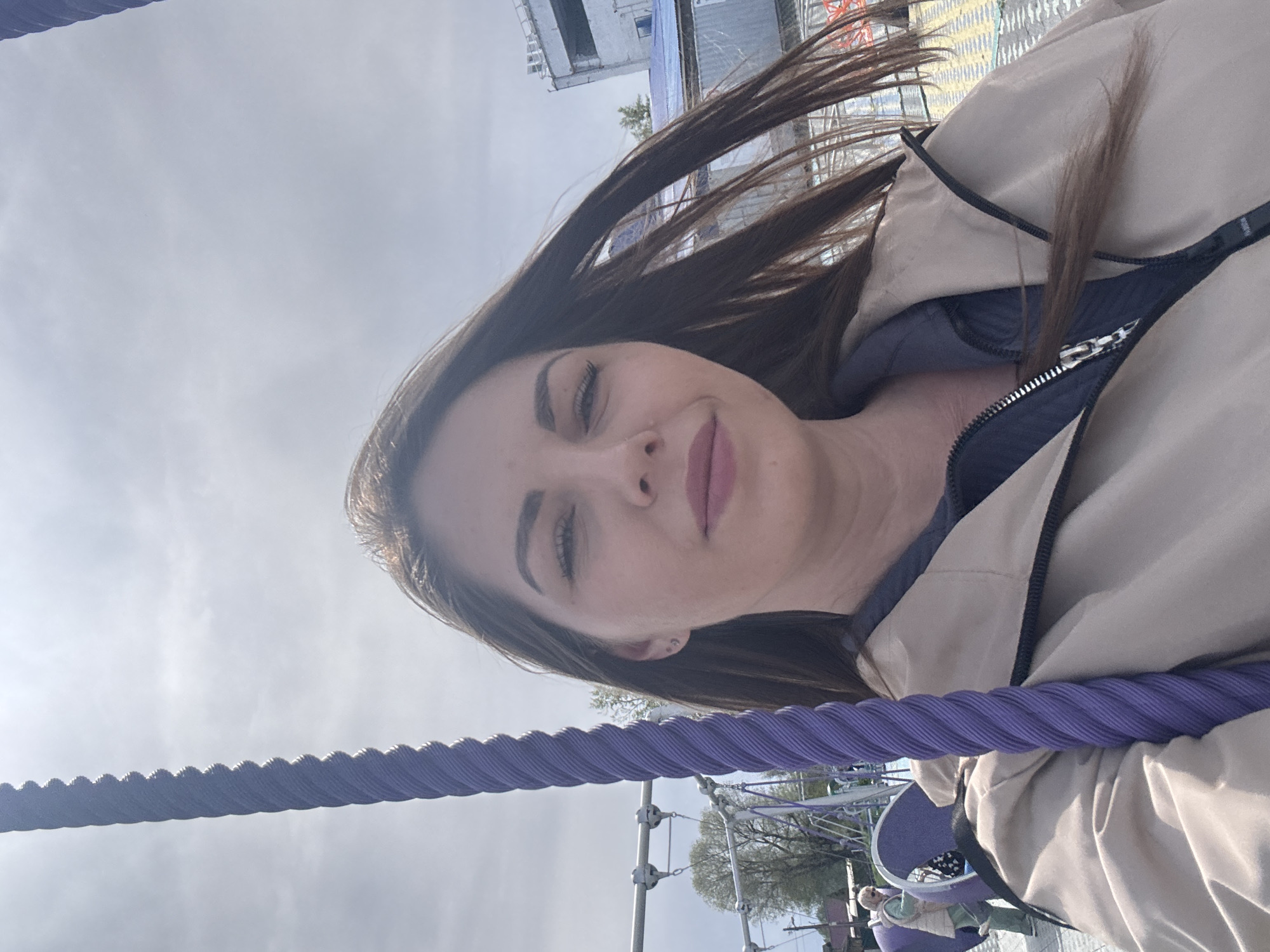 A woman poses for a selfie outdoors. She has brown hair and fair skin, and she is wearing a beige jacket. A thick, purple rope runs diagonally across the bottom of the frame. In the background, there is a metal playground structure and some trees. The image was taken in Синьково, Russia.
