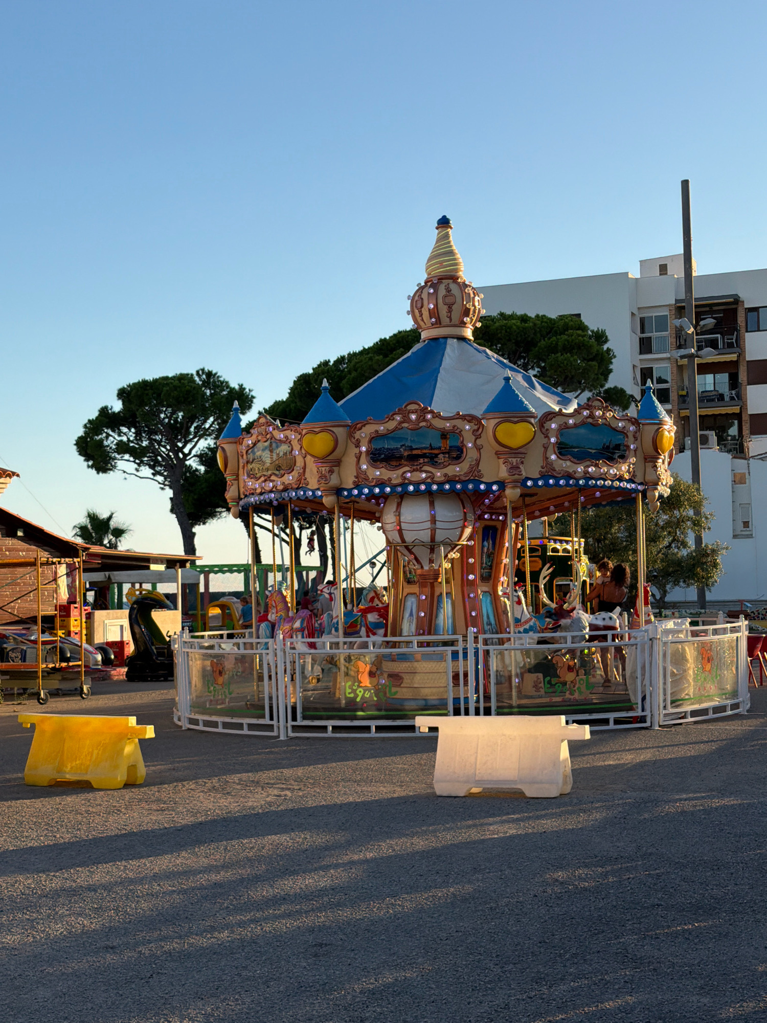 In Vilafortuny, Spain, a carousel is set up in an open, paved area. The carousel, adorned with lights, features various animals for children to ride, including horses and a deer. Several people are visible on and around the carousel, with some children actively riding. The scene is framed by trees and a multi-story building in the background, under a clear blue sky. In the foreground, two plastic barriers, one yellow and one white, sit on the ground, casting long shadows. The overall setting appears to be an amusement or fairground area.