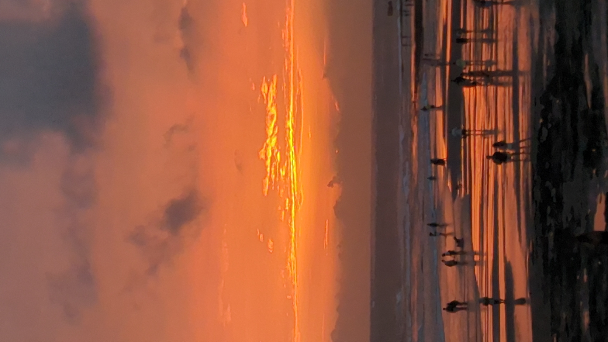 The image captures a serene sunset at the beach in North Kuta, Indonesia. The sky is ablaze with hues of orange and yellow, with dark clouds interspersed throughout. The sun, a bright streak of light, is low on the horizon, casting long shadows across the wet sand. Several people are seen scattered across the beach, some walking individually, others in pairs, and a few in small groups. They appear to be enjoying the evening, with their silhouettes reflecting on the water-covered sand. The ocean stretches out in the background, with gentle waves lapping at the shore. A pier or boardwalk is faintly visible on the right side of the image. The overall atmosphere is peaceful and contemplative, characteristic of a sunset gathering on a beach.
