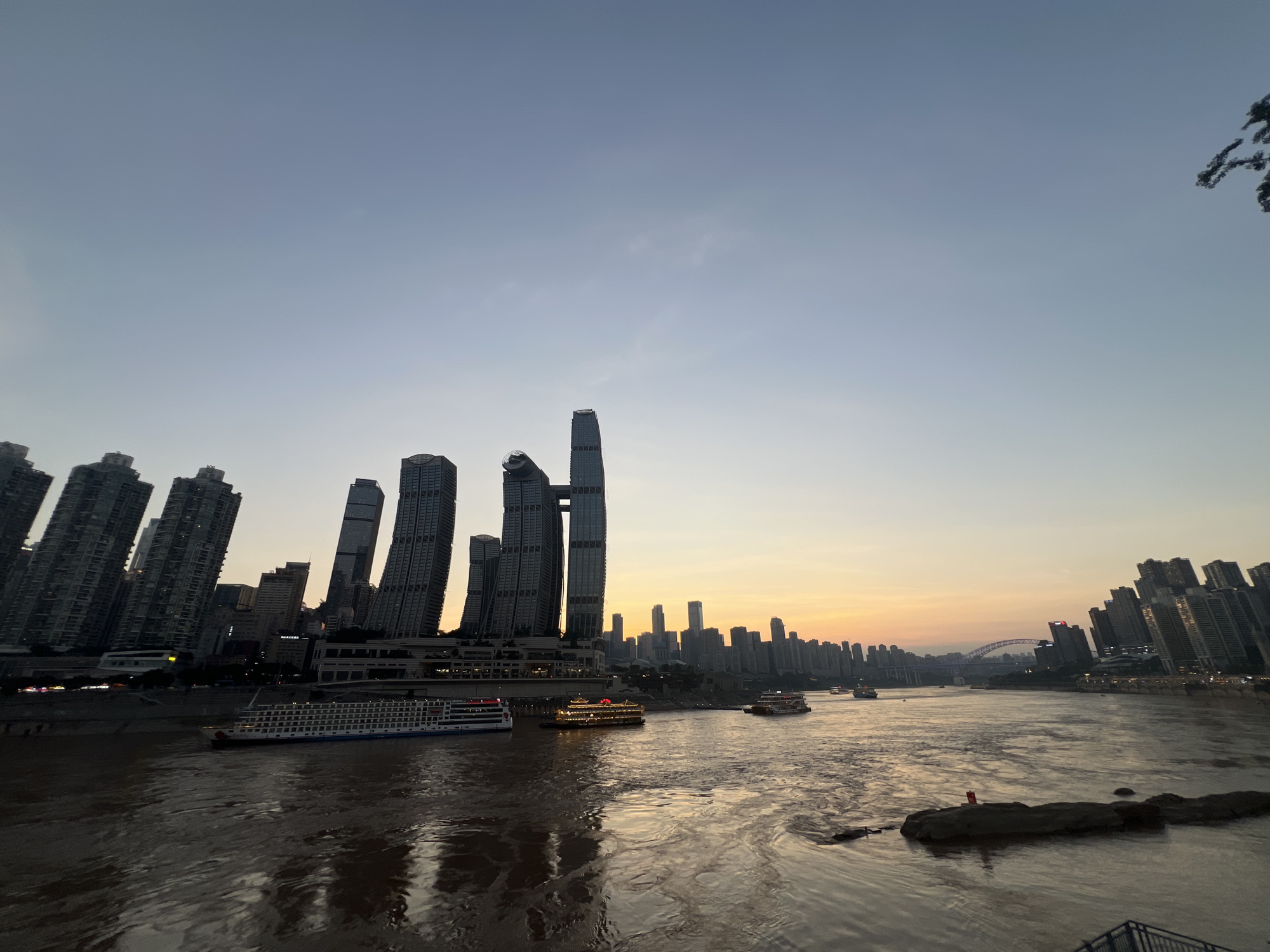The image depicts a wide river flowing through a city skyline at dusk. Several large, modern buildings, including skyscrapers and apartment complexes, line the riverbanks. The sky is a gradient of light blue and orange, indicating the sunset. Several boats, including a large passenger ship with multiple decks and smaller tour boats, are visible on the river. The water is a murky brown and reflects the city lights and the sky. In the foreground, a few rocks are partially submerged in the water, with one rock having a small red object on it. The scene is observed from a slightly elevated perspective, suggesting the photographer is on a bridge or a higher vantage point overlooking the river and the Nan'an District of Chongqing, China.